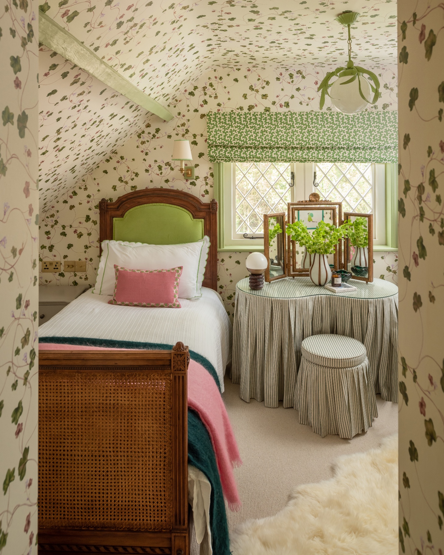 an eaves bedroom with cream and green trailing ivy wallpaper, a single dark wooden bed with white bedding, a pink cushion and a pink and teal bed spread and a dressing table with a white and grey stripe fabric skirt and matching stool
