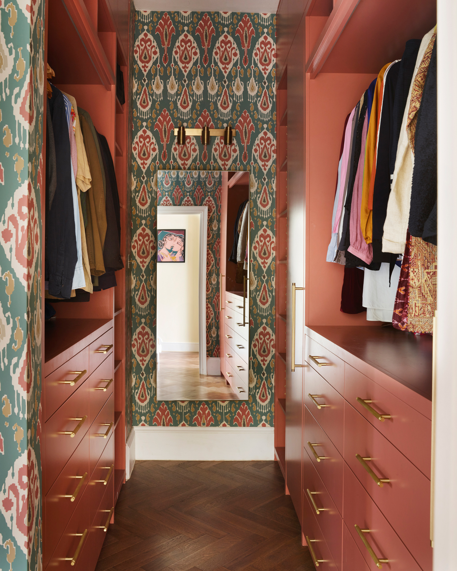 a walk in wardrobe with orange/red painted cabinets, open wardrobes filled with clothes and a back wall covered in a green and red ikat print with a full length mirror