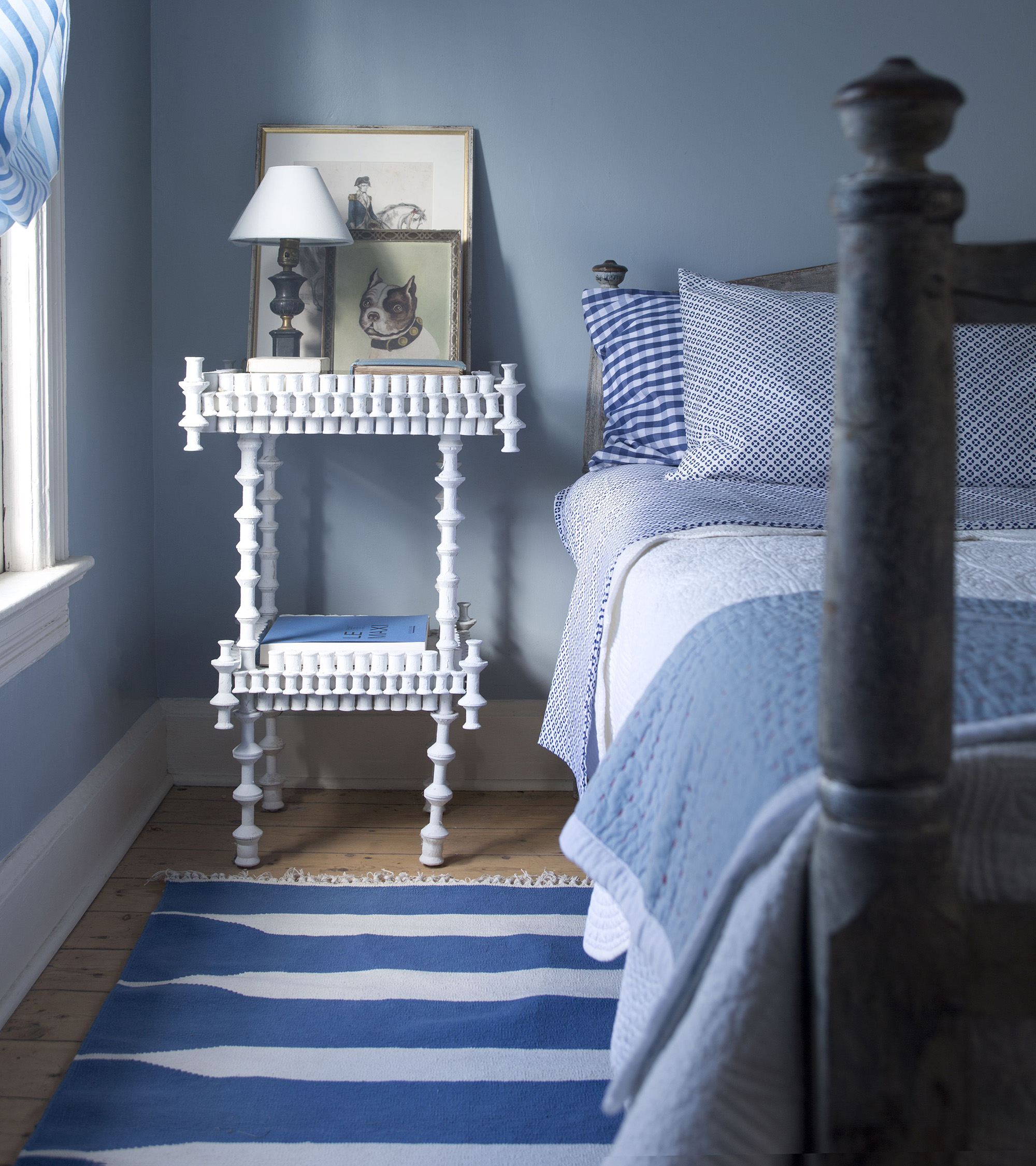 a blue bedroom with a blue adn white striped rug, a white bedside table with two prints and a lamp on it, and a bed with blue and white bedding