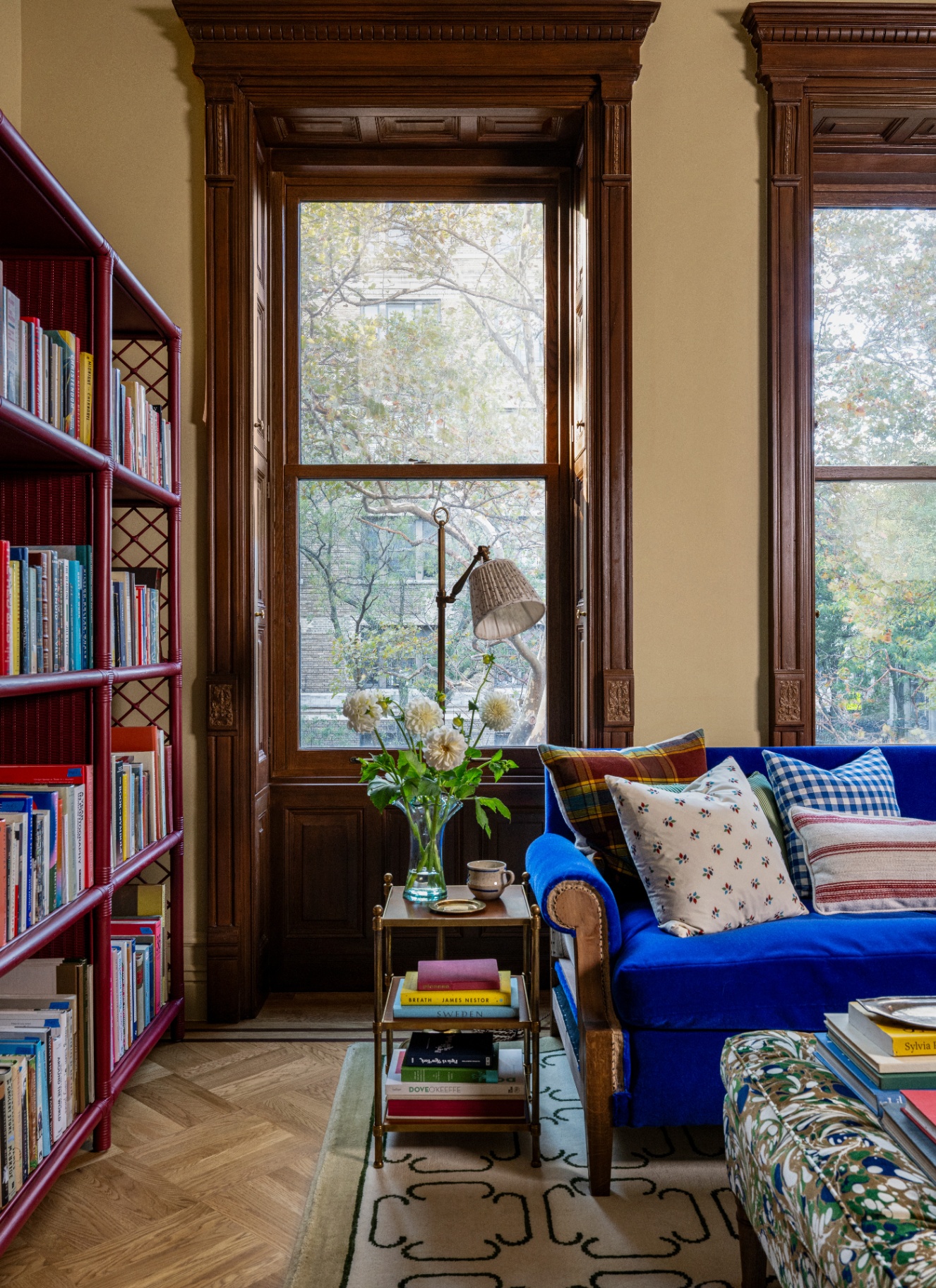 A sunlit room featuring rich dark timber window framing, dark cane-style bookshelves, a vibrant royal blue velvet sofa, and layered patterned textiles