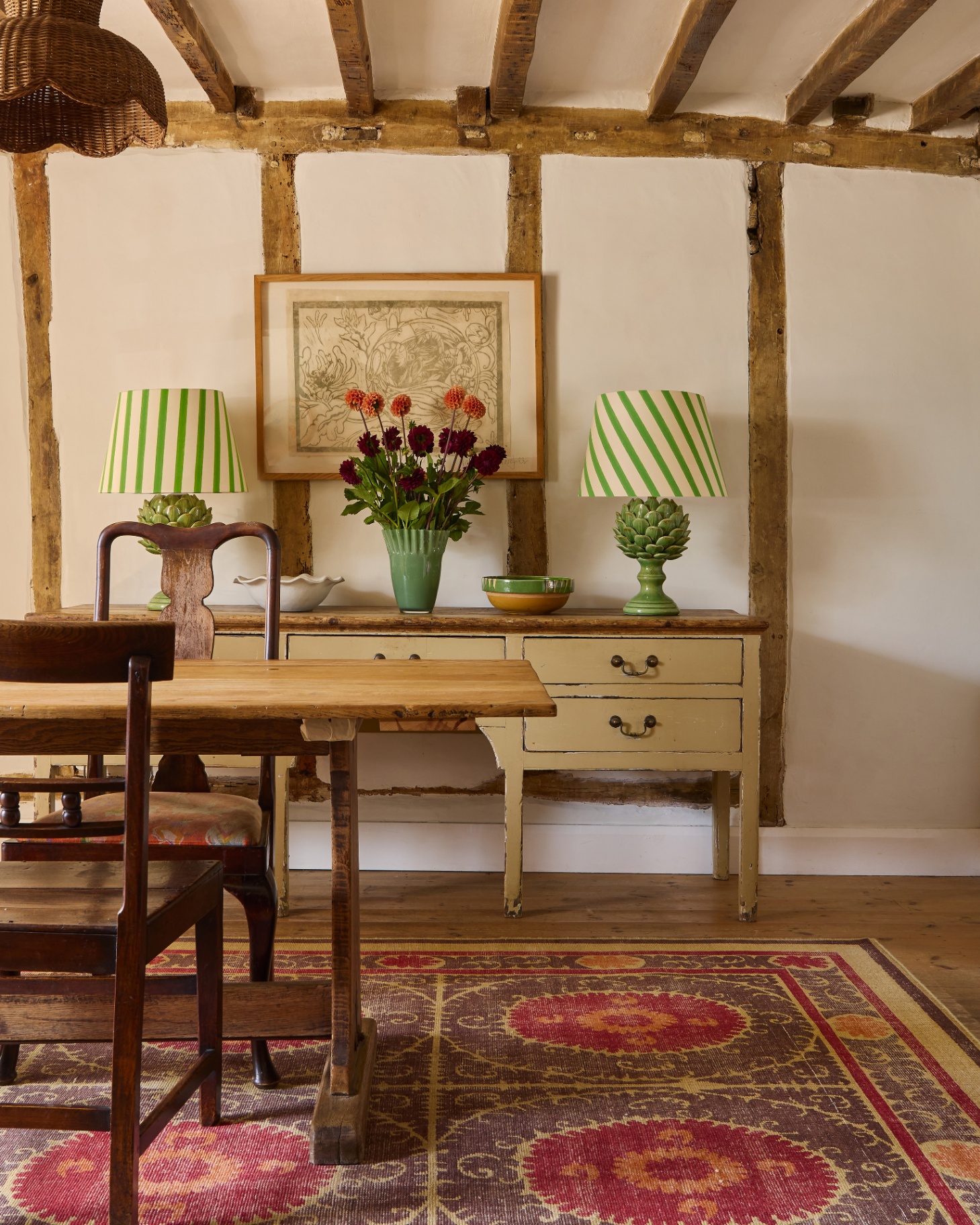 a dining room with a dark red, beige and purple patterned rug under a wooden dining table and chairs with a wooden cream painted sideboard with two green artichoke lamps