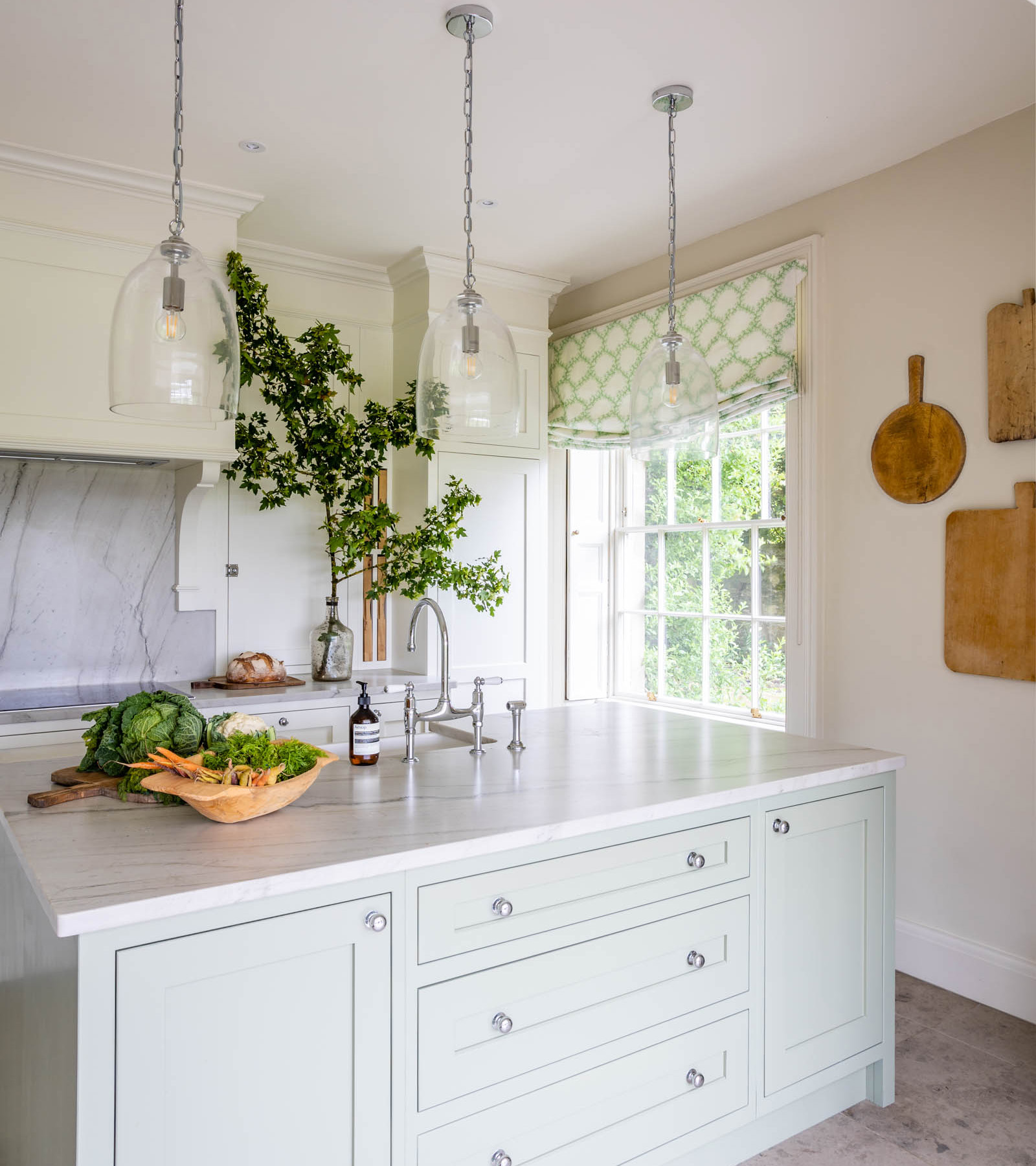 a kitchen with white walls and wooden chopping boards hanging on the wall, a sage green kitchen island with white marble top and three clear glass pendant hanging above