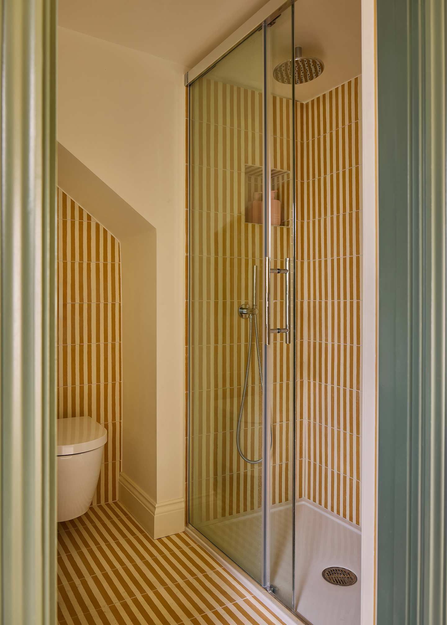 a shower room with striped wall and floor tiles. There is an enclosed shower with silver hardware and a floating toilet