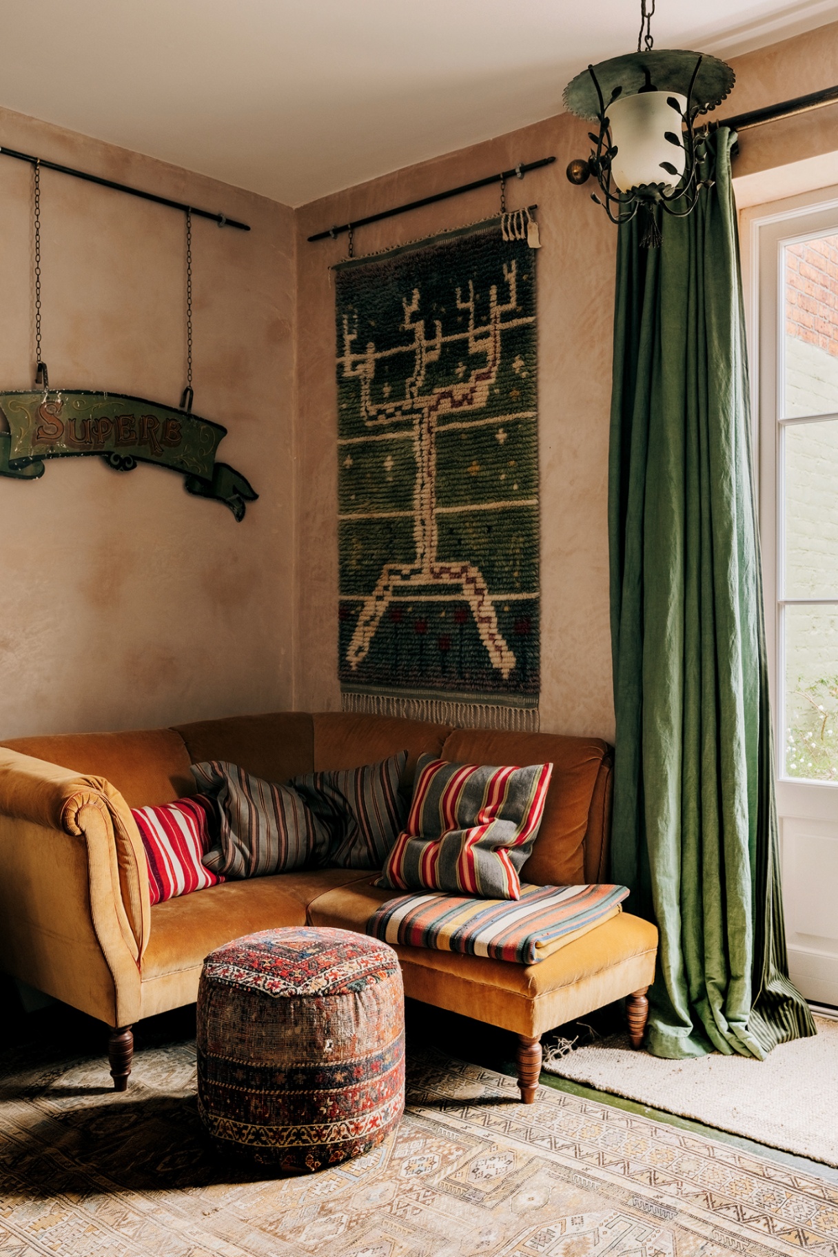 the corner of a living room with a compact corner sofa in orange velvet, a patterned round footstool, a long green curtain hangs to the right and on the wall hangs a green and white rug and a vintage sign