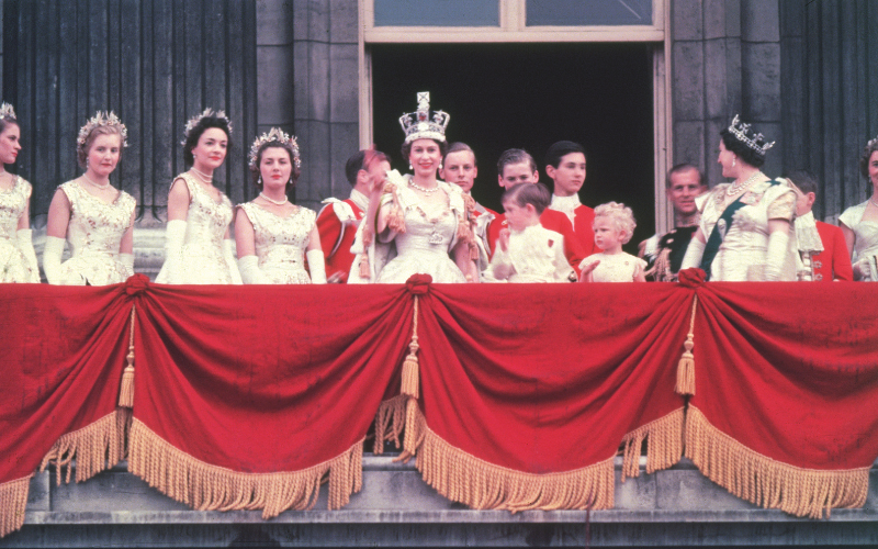 The newly crowned Queen Elizabeth II waves to the crowd from the balcony at Buckingham Palace with her children next to her, she wears a white gown and robe with a crown and the balcony is draped in red curtains