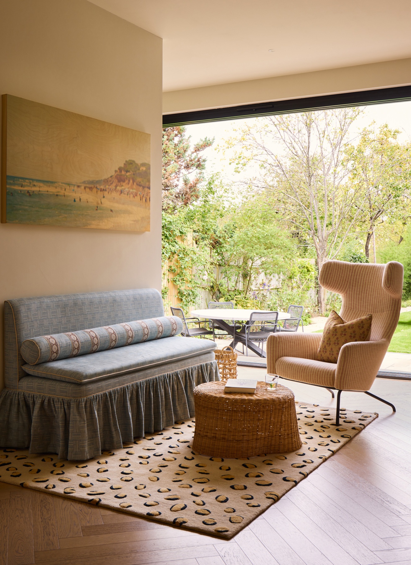 a sitting room with a pale blue skirted sofa, a small rattan coffee table, a modern striped armchair and an open french door looking out onto the garden