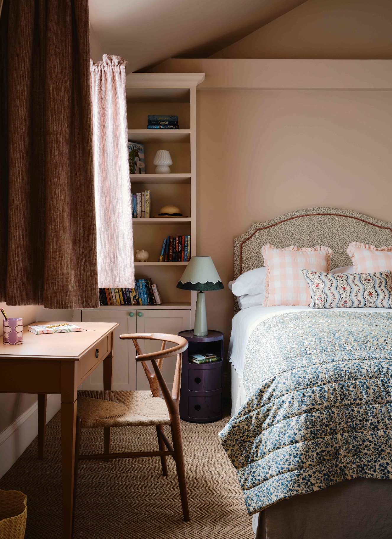 a bedroom with pale pink walls, a small wooden desk and chair facing the window and a bed with a patterned headboard, pink gingham cushions and a blue and white floral bed spread