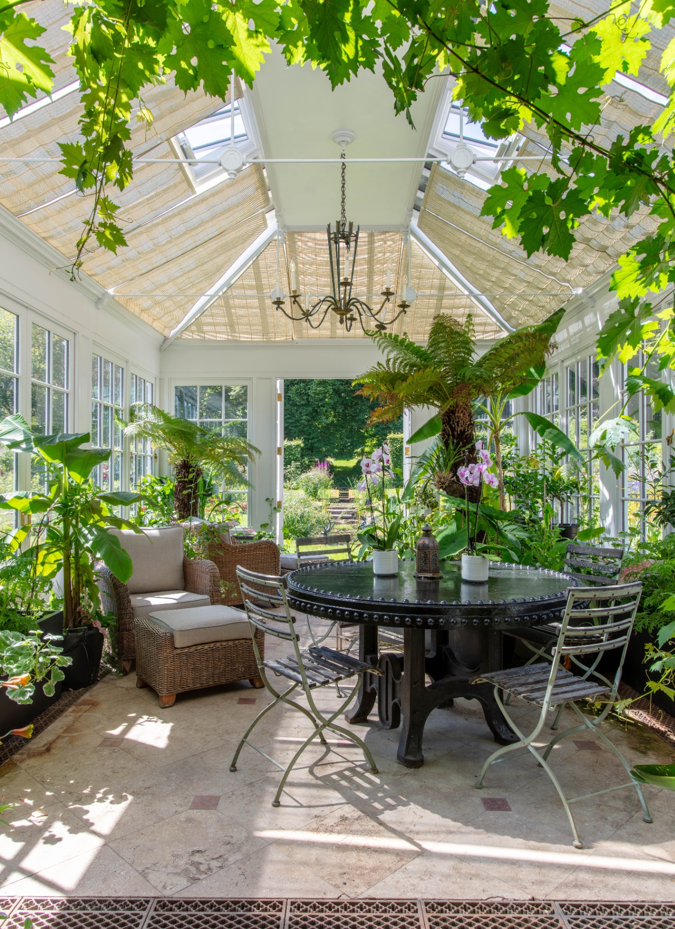 a large garden room conservatory with large green plants, cream stone flooring and a large ground table with folding bistro chairs
