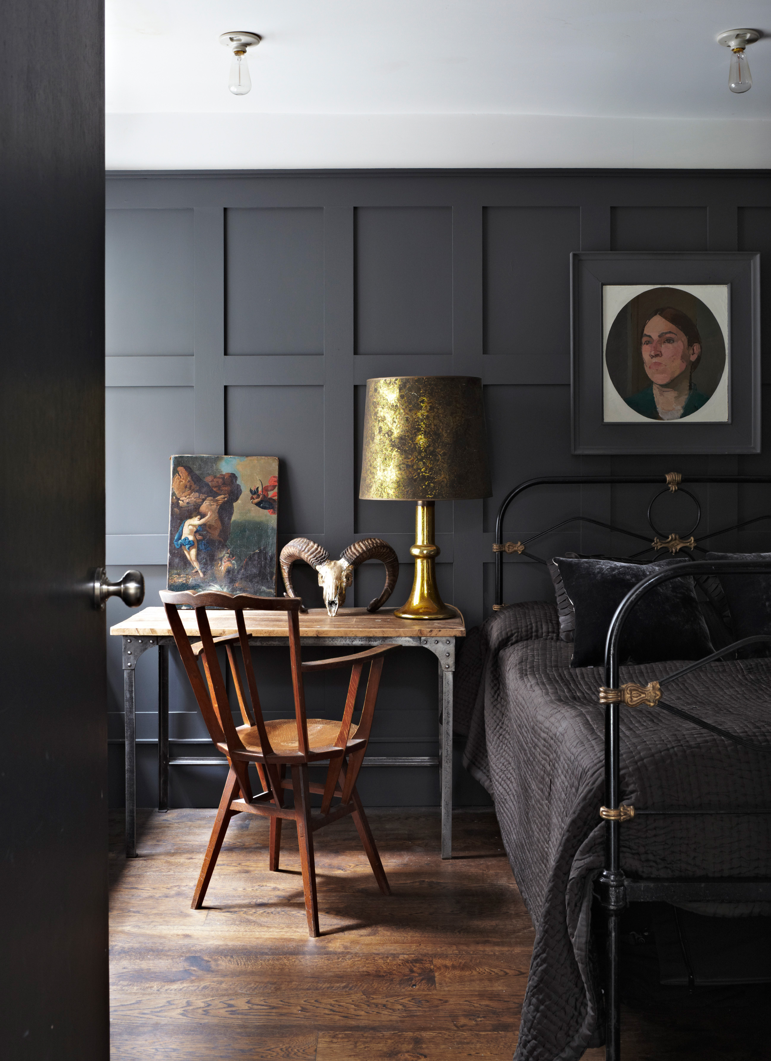 Charcoal grey grid paneled bedroom wall behind a black iron bed frame, styled with a rustic wooden desk, brass table lamp, and framed artwork