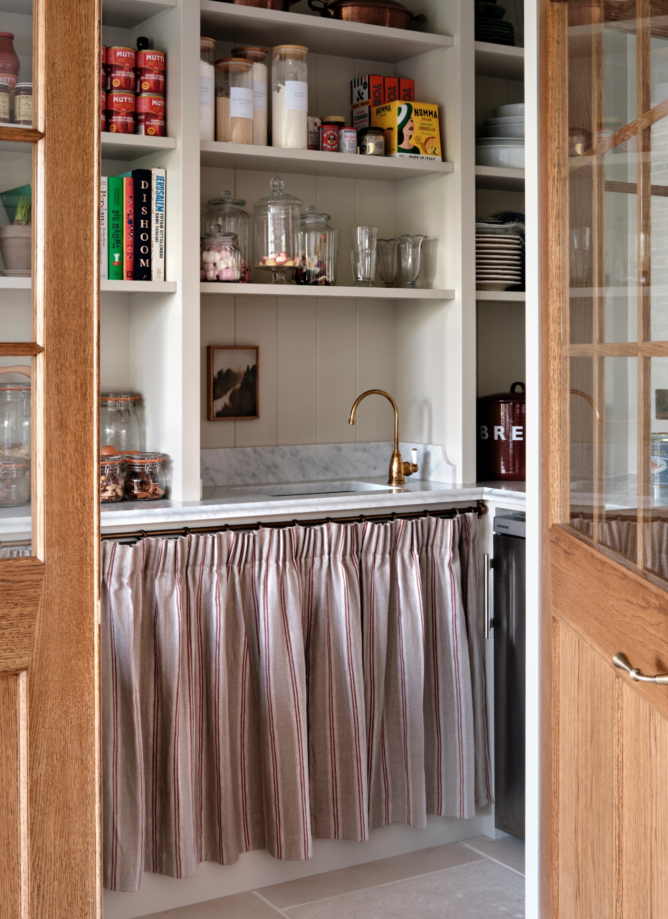 a pantry with white marble counterstops, a grey and red stripe skirted cupboard and shelves filled with jars and tins of food