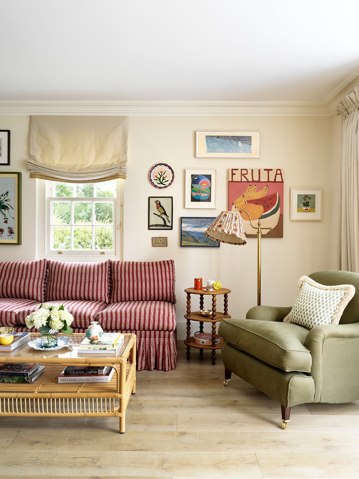 A seating area in a kitchen with a red striped sofa, green armchair, brass floor lamp with pleated shade, bobbin side table and coffee table. Above a collection of art hangs on the neutral walls