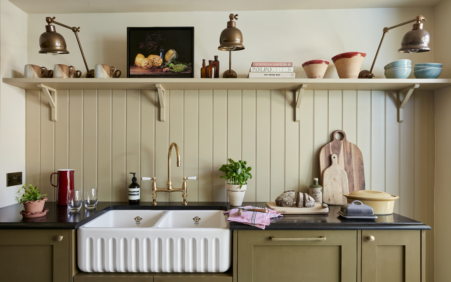 a kitchen with olive green cabinetry, cream tongue and groove panelled walls and a long open shelf filled with cups, books and bowls