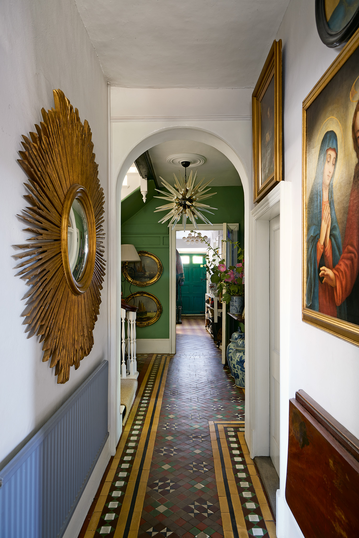 a narrow hallway with an arched doorway at the end, the flooring is patterned, colourful victorian-style tiles and on the walls are large religious artworks and a gold starburst mirror