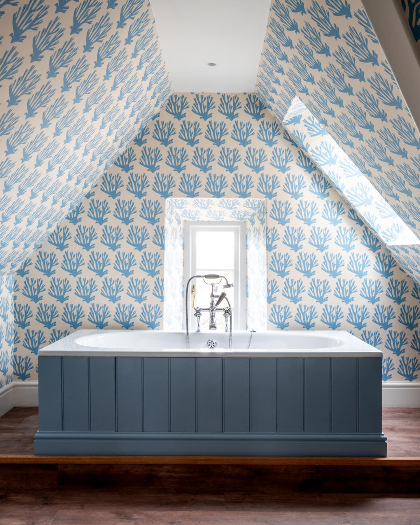 a bathroom with sloping ceilings covered in a blue and white coral block printed wallpaper and a blue paneled freestanding bath
