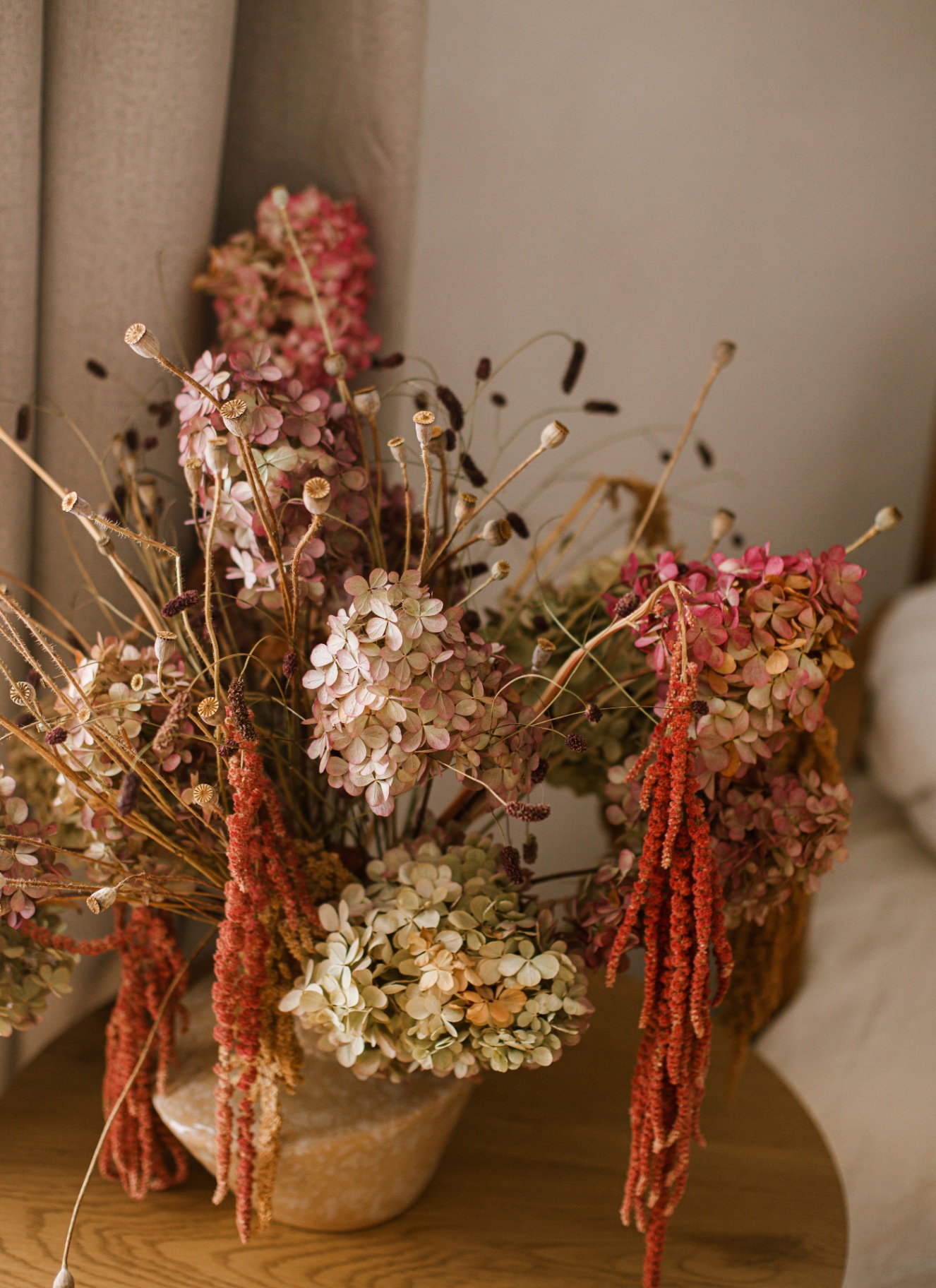 A rich, rustic bouquet of dried mophead hydrangeas, poppy seed heads, and cascading red amaranth in an earthy ceramic vase on a wooden table, with natural linen curtains in the background