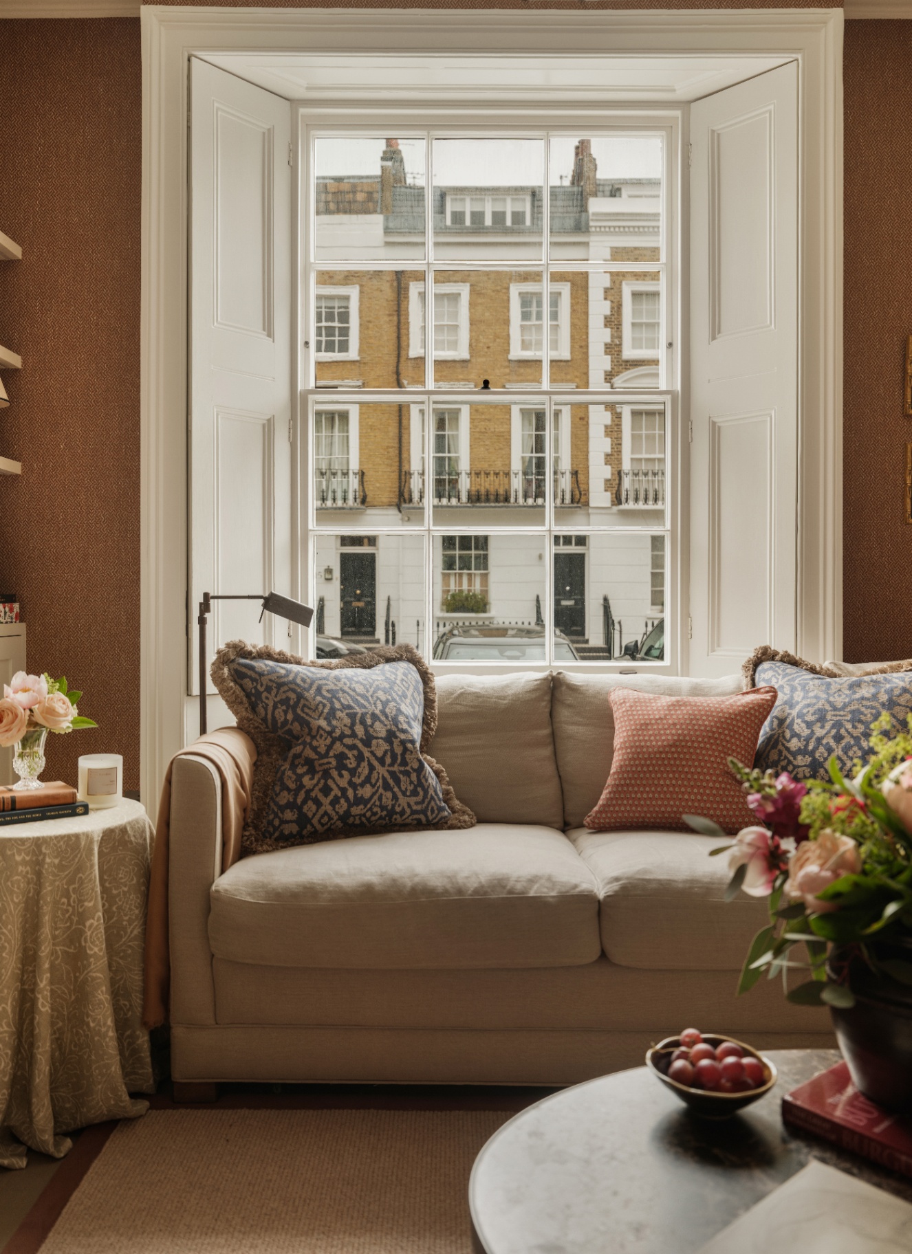 Light beige couch layered with printed cushions positioned before a large sash window with white indoor shutters. Warm brown wallpaper, fresh flowers, and a draped side table complete the refined setting