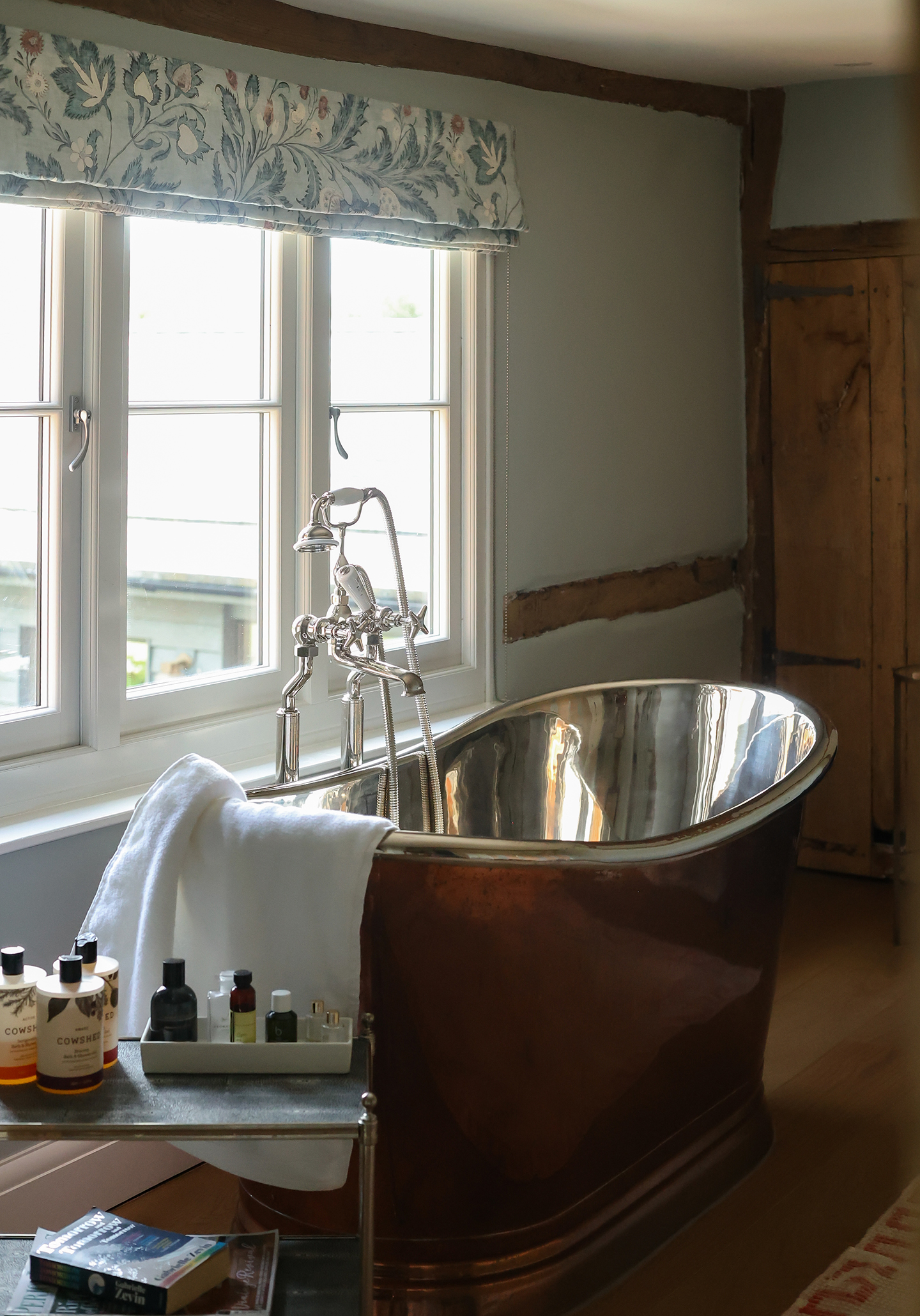A bathroom with a copper freestanding bath with a nickel interior set in front of a window dressed with a blue floral blind. The walls are pale blue and wooden beams are set within the walls
