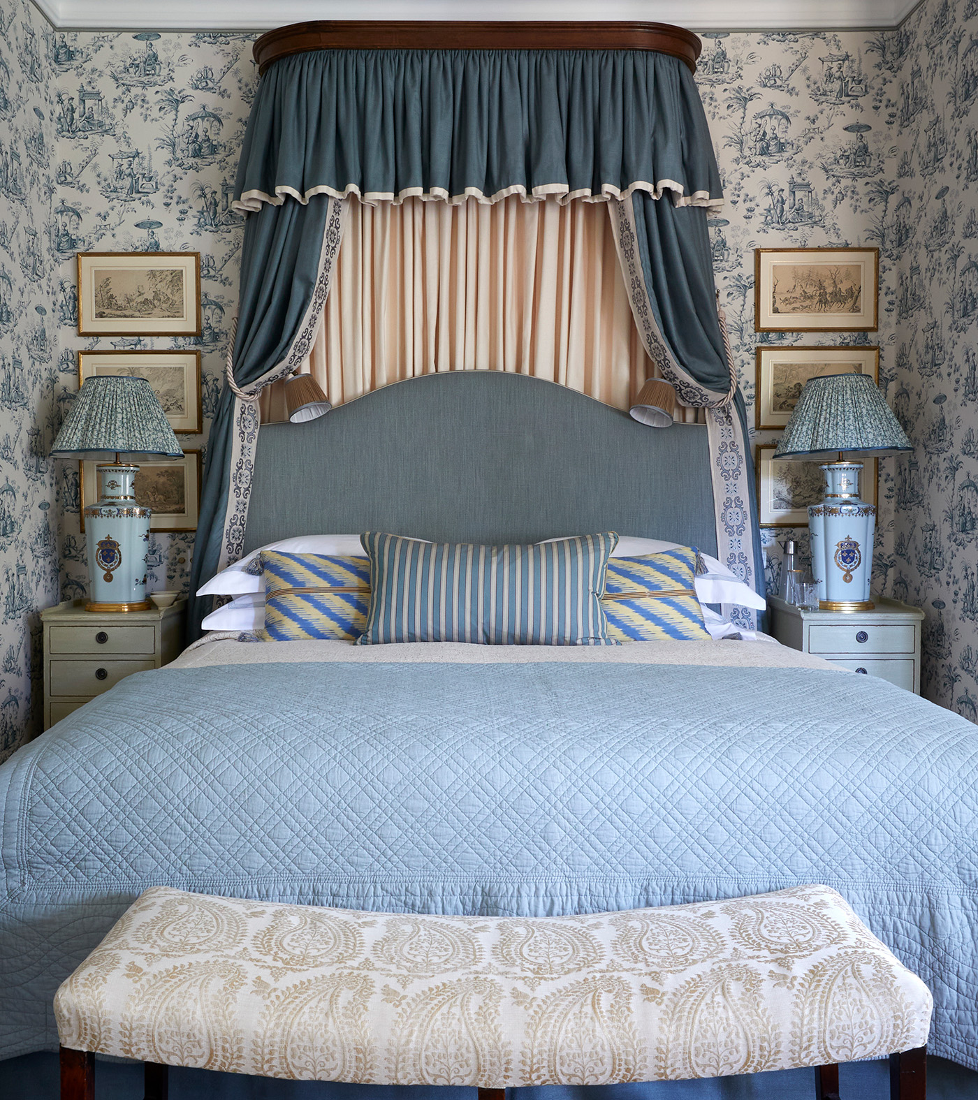 a traditional blue bedroom scheme featuring a double bed with a canopy and blue bedding, blue and white patterned wallpaper, two bedside tables with lamps and art on the wall and an upholstered bench at the end of the bed