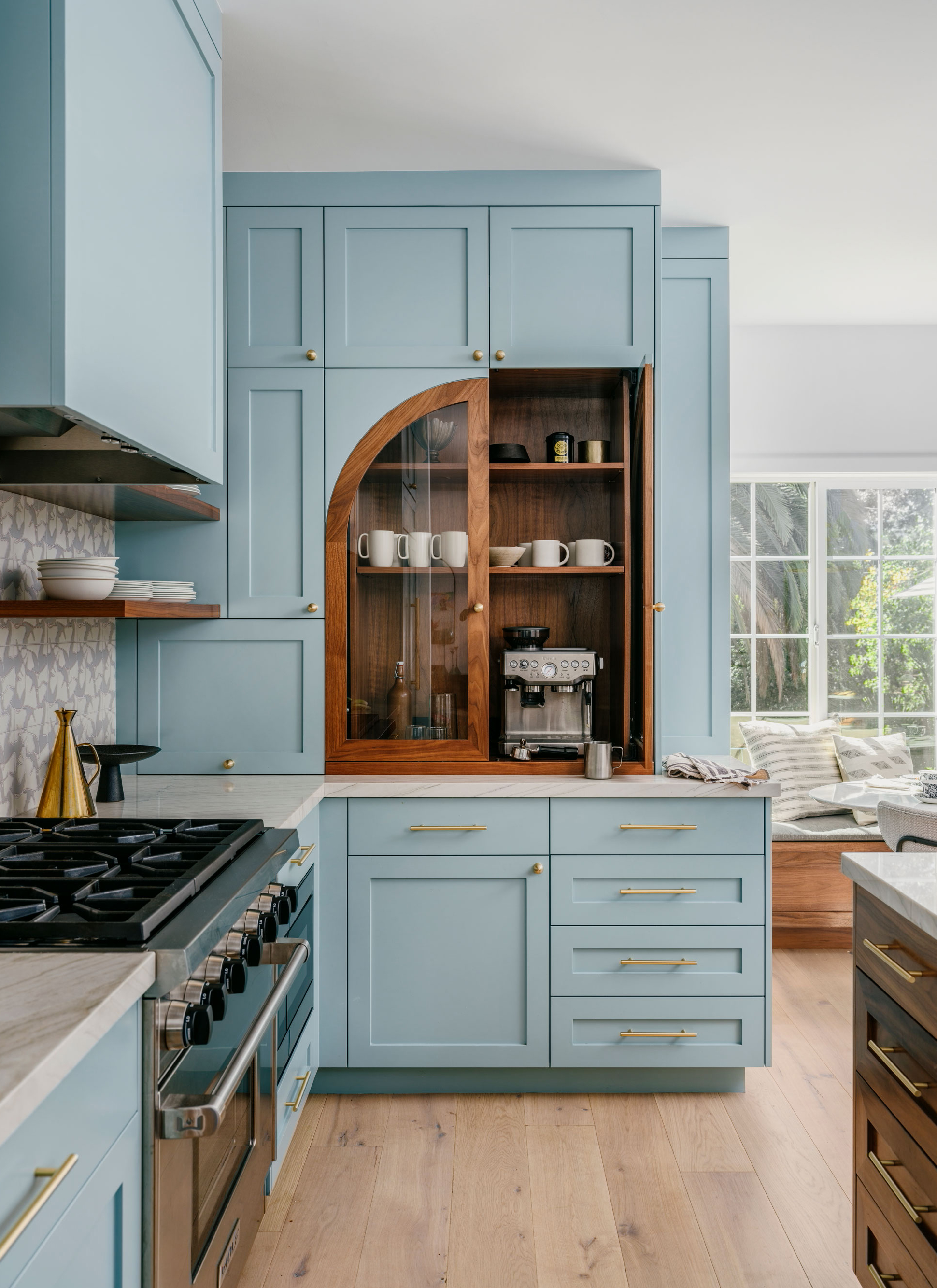 Light blue kitchen cabinetry featuring a built-in coffee station framed by an arched walnut glazed door with an espresso machine and mug shelves inside.