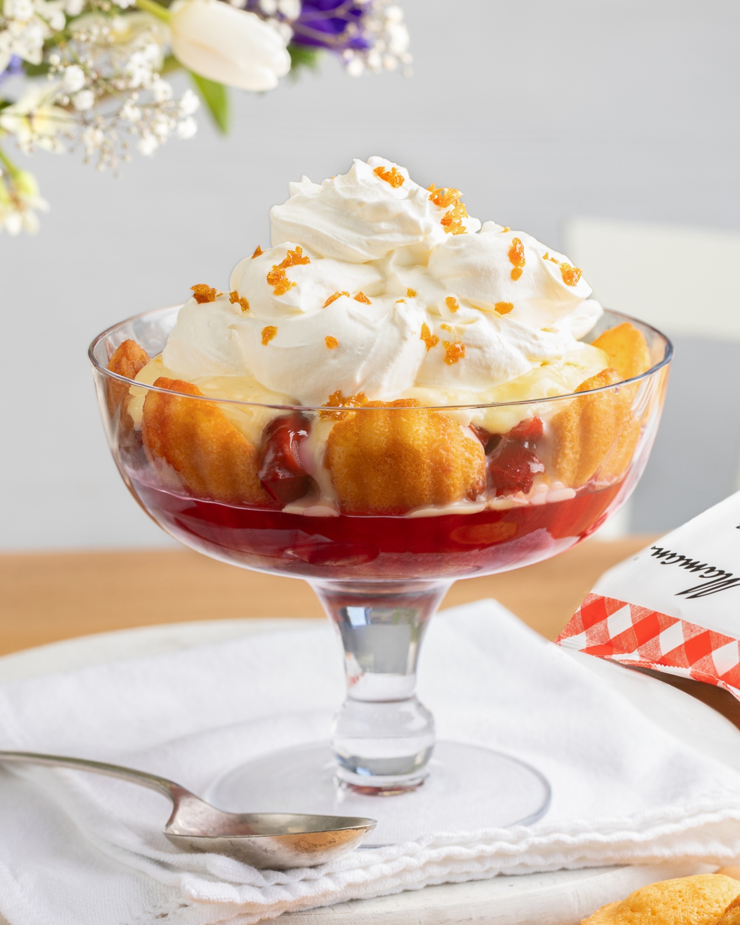 a plum and madeline trifle in a glass footed bowl on a white tablecloth with a dessert spoon
