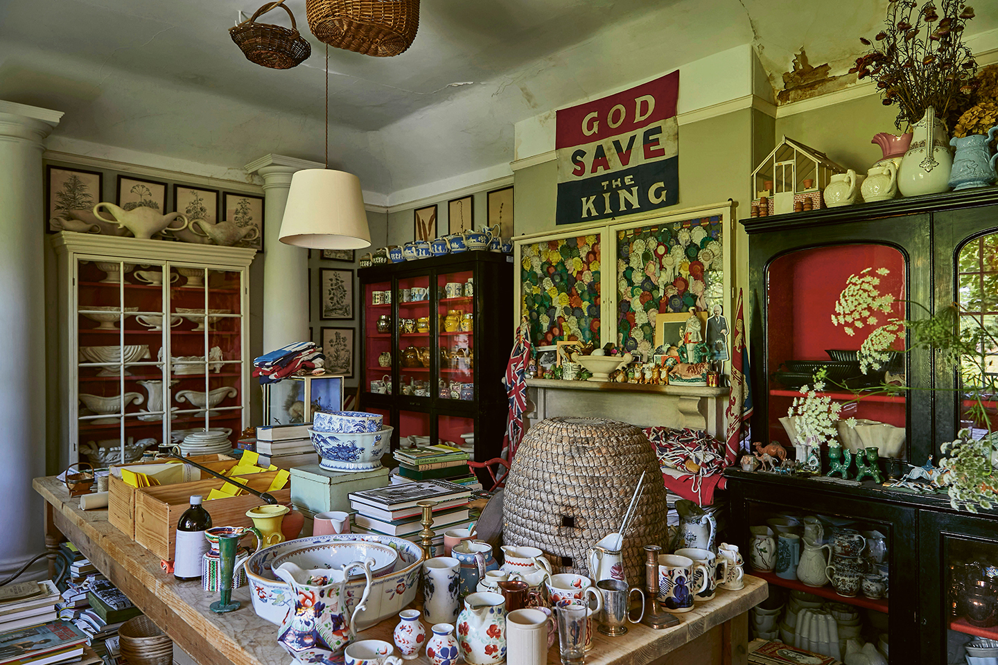 a flower room filled with cabinets and a large central table all filled with china, seed packets and antique objects. A pendant light and two wicker baskets hang from the middle of the ceiling 
