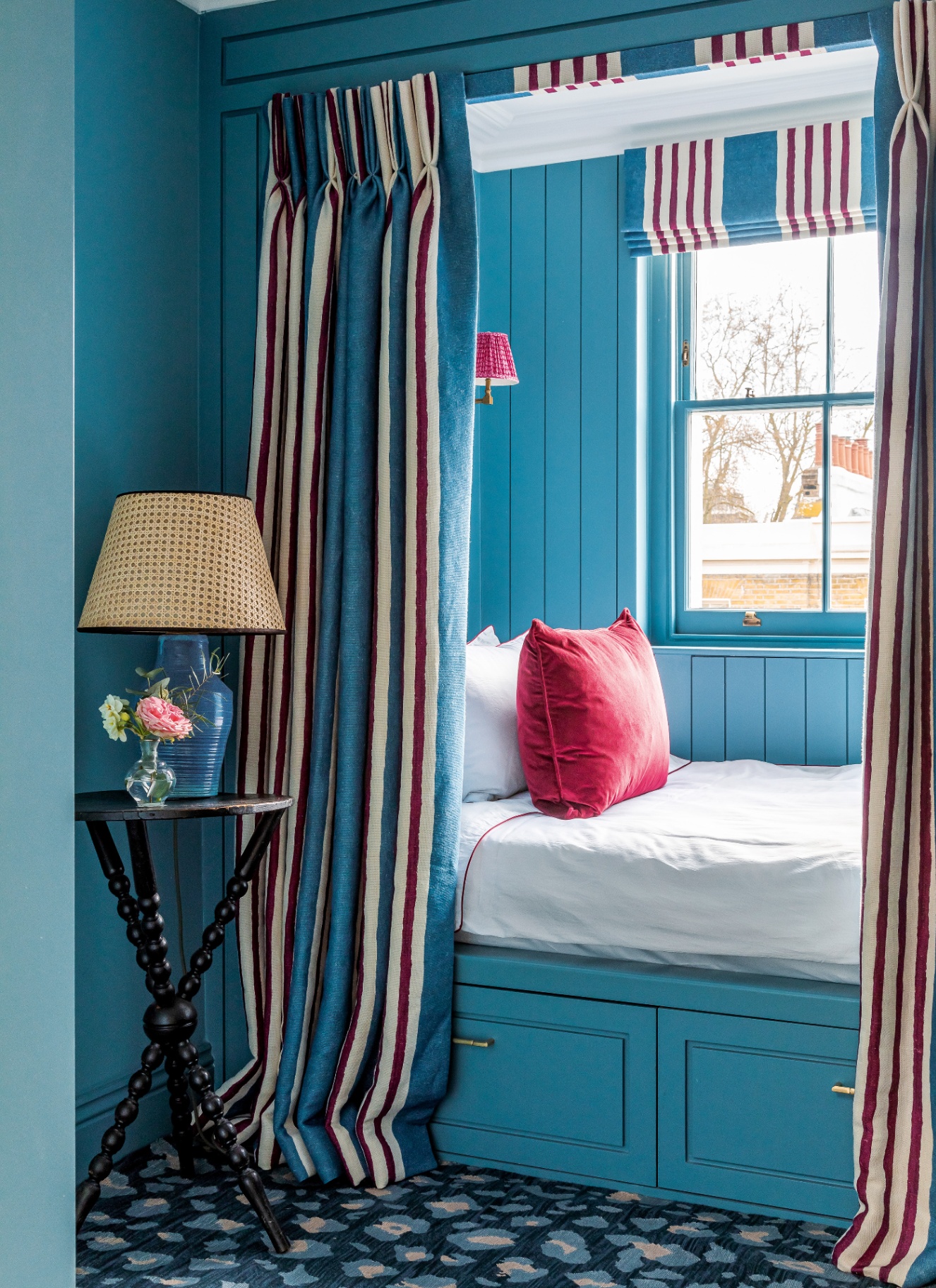 a bed nook painted in bright blue with blue, red and white curtains covering it and matching blind on the window