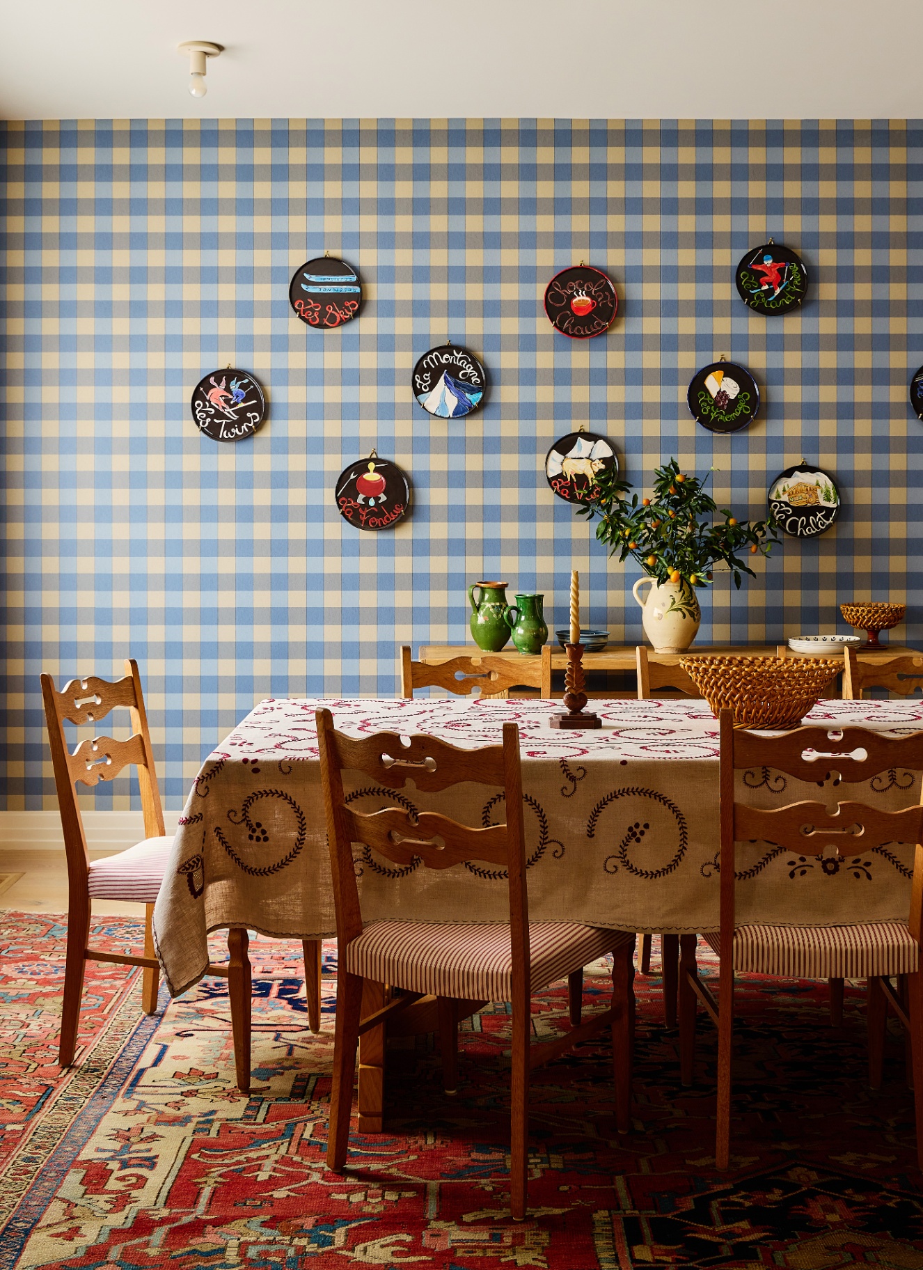 Rustic dining space with blue and beige gingham wallpaper, decorative plate display, wooden table with embroidered cloth, and a red oriental rug