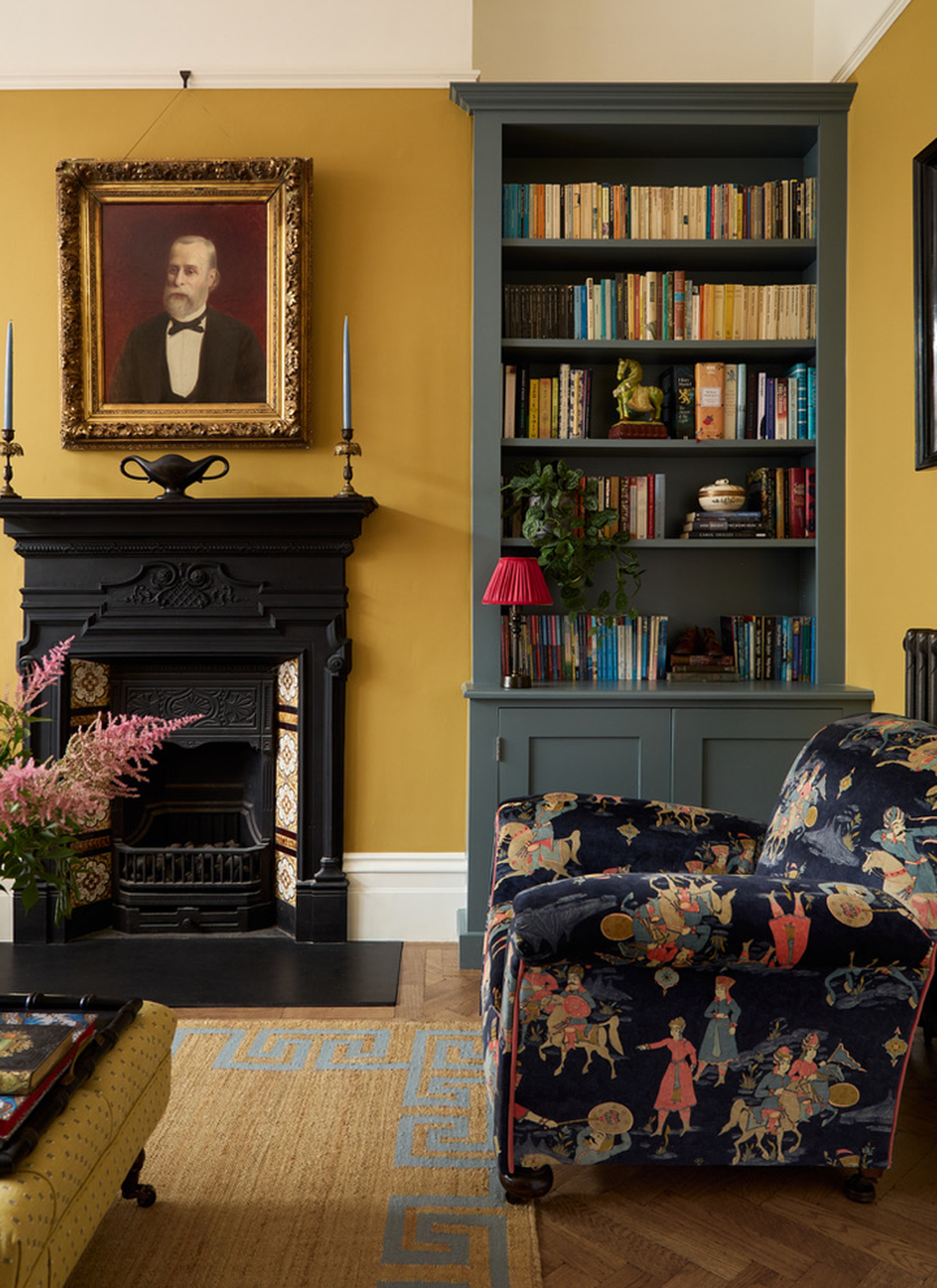 A vibrant reading nook with ochre yellow walls, a black fireplace surround, dark grey built-in bookshelves, and an armchair upholstered in a bold navy equestrian print