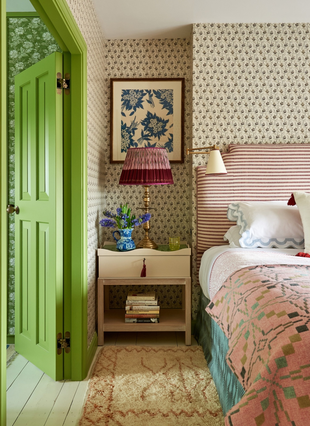a bedroom with a green open door, a bed with a pink headboard and a pink patterned bedspread and a cream bedshire table with a red plated lamp