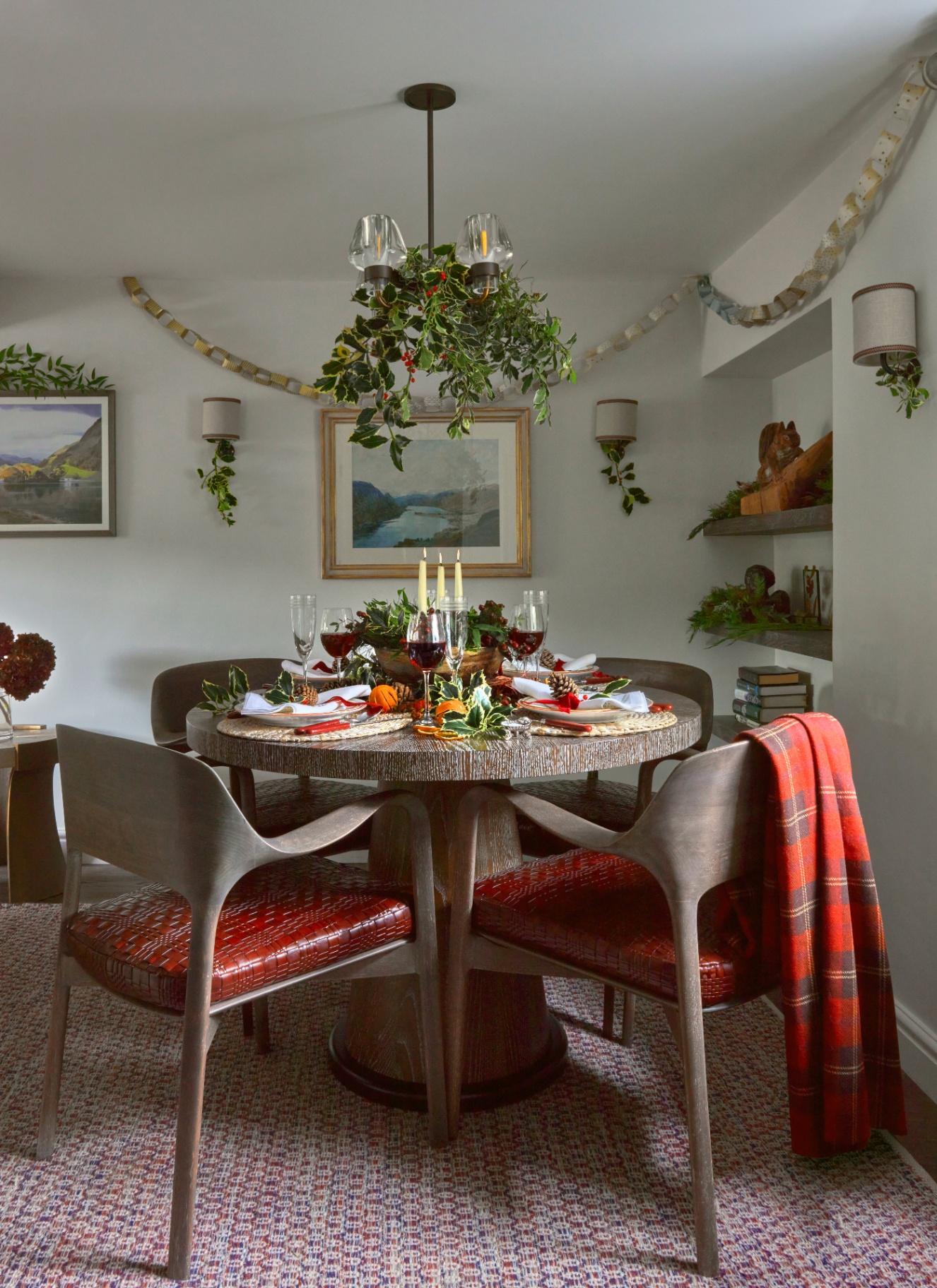 A round dining table centered with a wooden bowl containing dried citrus, holly, and tall candles under a light decorated with winter greens.