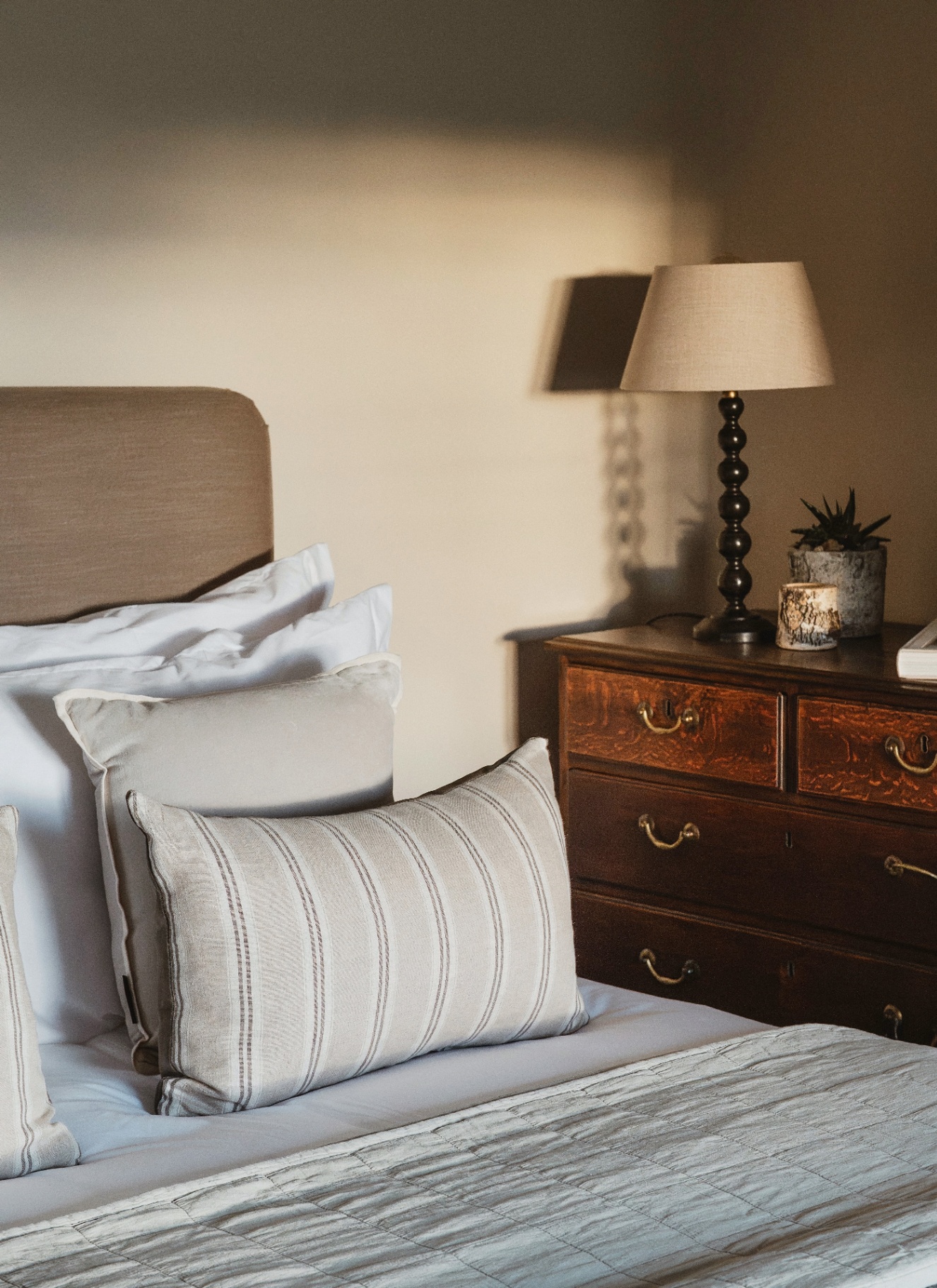 a bedroom with a bed with a beige headboard and striped cushions and a dark wooden chest of drawers with a cream lamp