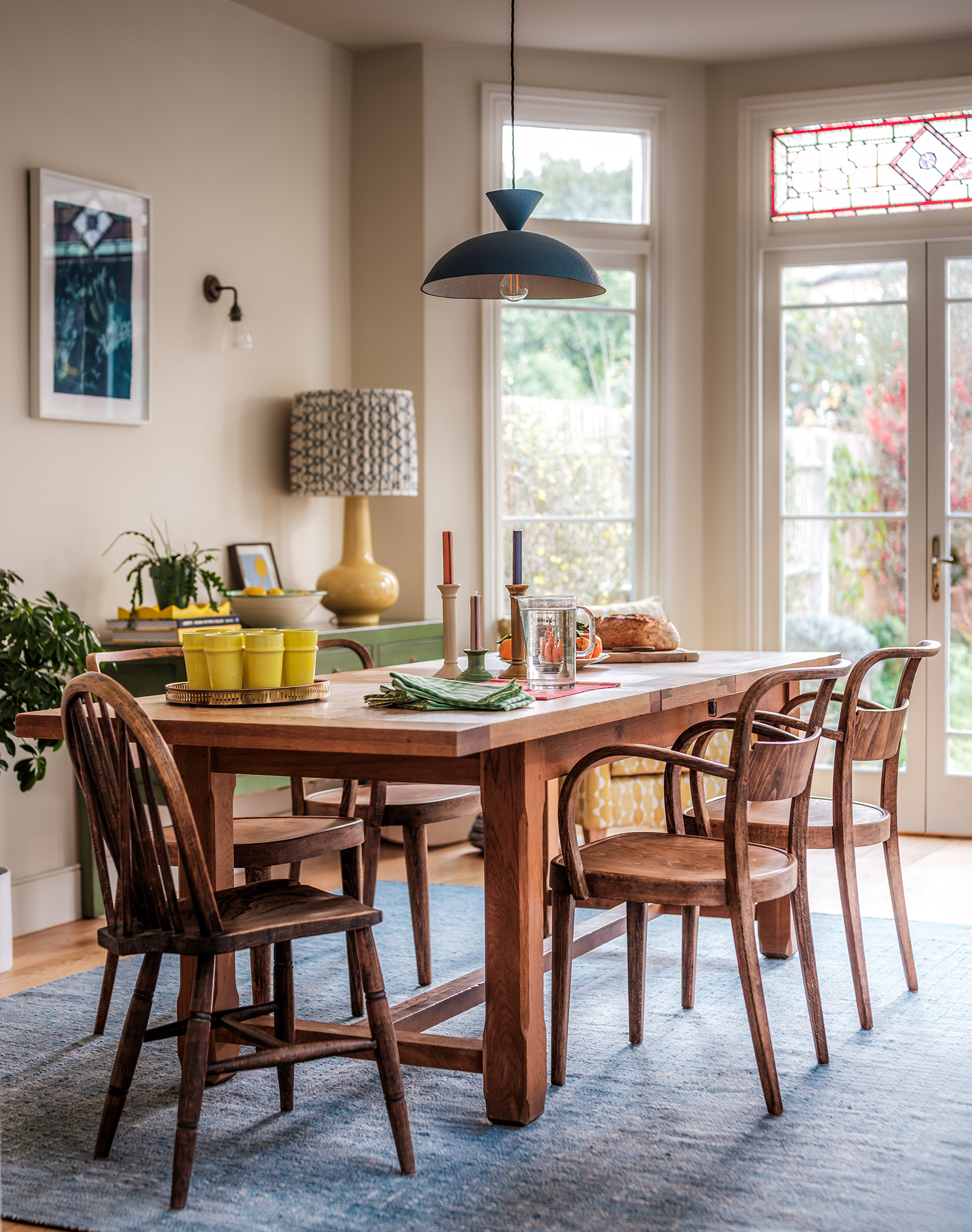 A large wooden dining table and mixed set of wooden chairs sit on a large blue rug with a blue pendant light above, in the background a partly-blurred green sideboard and a yellow table lamp with a pleated fabric shade can be seen next to some french doors