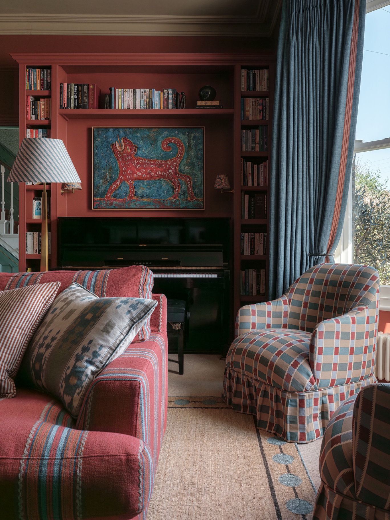 A cosy snug with dark red walls and tall blue curtains dressing the bay window. In the background a piano is surrounded by built-in bookcases and a vibrant artwork. In the foreground sits a striped sofa and pair of patterned armchairs 