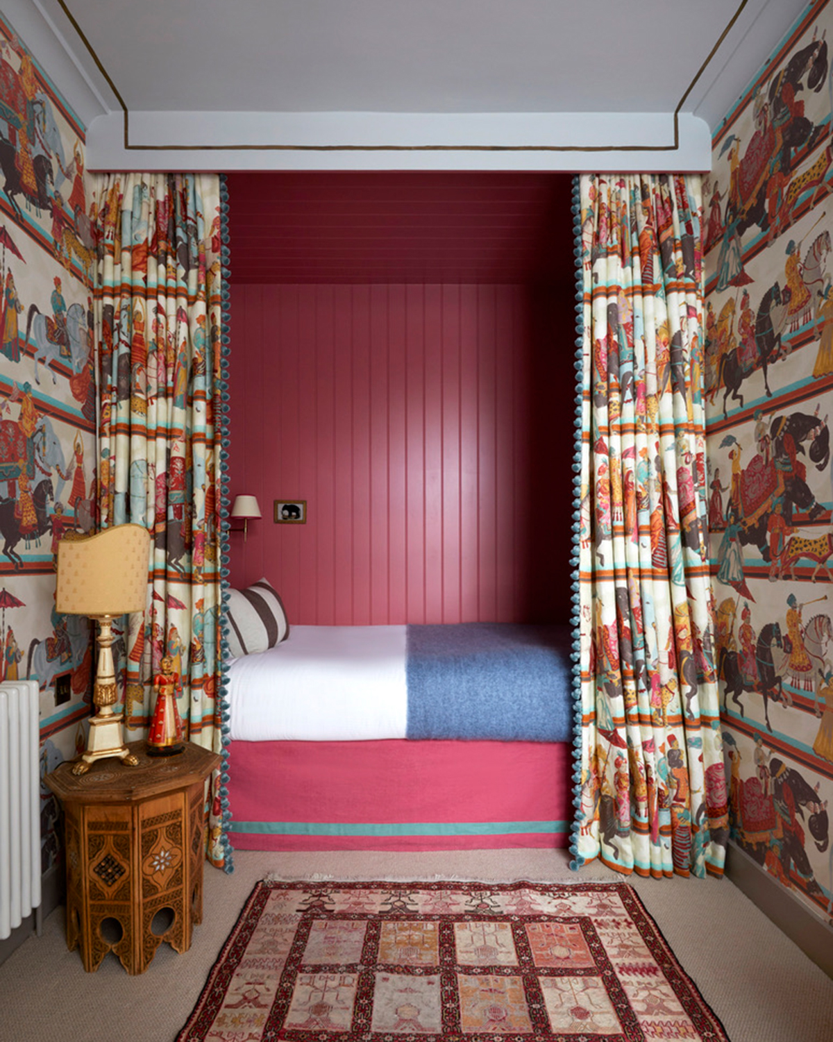 a small bedroom with white, red and teal patterned wallpaper and matching bed curtain, a bed alcove with dark red panelling, a dark wood bedside table with a large cream and white lamp and a dark red persian rug