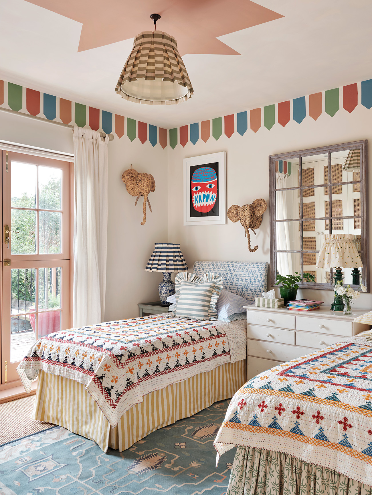 a childrens bedroom with cream walls with mulitcoloured painted bunting on the walls and twin beds with yellow and white stripe valances