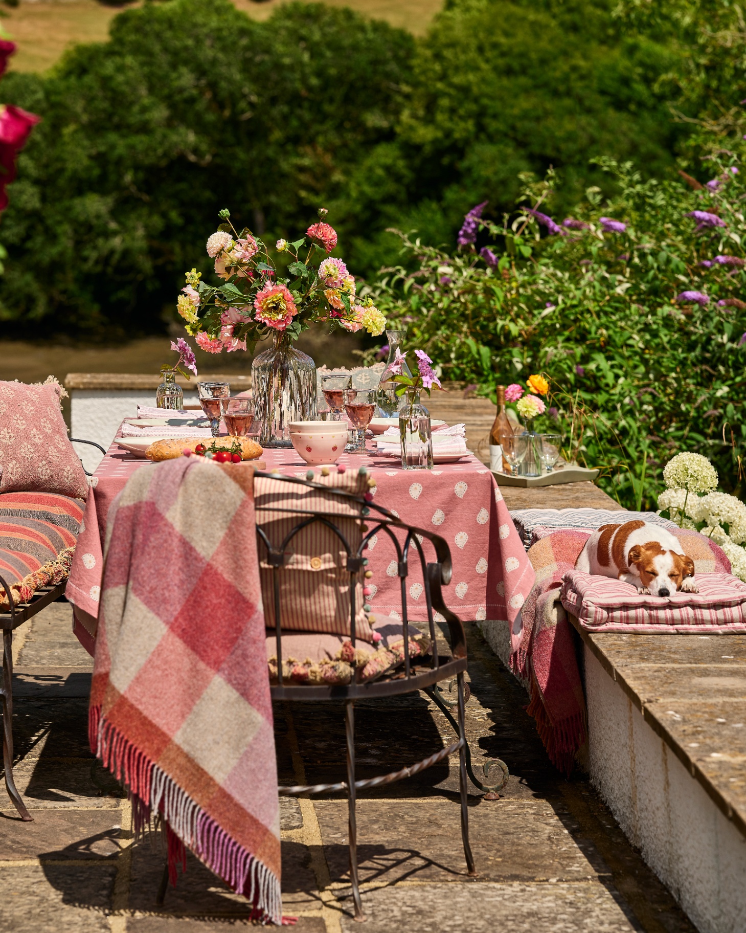 outdoor table with pink patterned throws hanging on the seats, a jack russel sleeping on a cushion on a wall a pink and white patterned tablecloth and a large glass vase full of flowers
