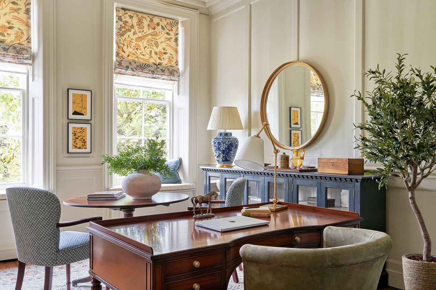 A home office with panelled walls painted off white, with a wooden desk and upholstered curved armchair. Behind sits a small circular table and two dining chairs. A long, glass-paned sideboard sits against the far wall with a circular mirror hanging above