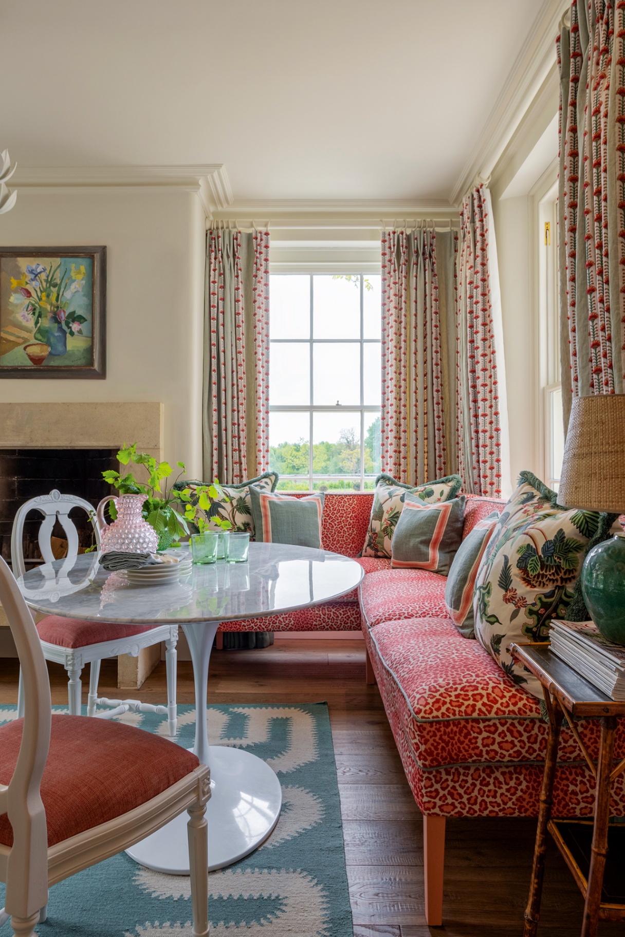 An oval marble table on a light blue and white rug is paired with an orange-pink leopard print banquette dressed with decorative cushions and two upholstered dining chairs. Patterned blue and red curtains line the windows behind the banquette