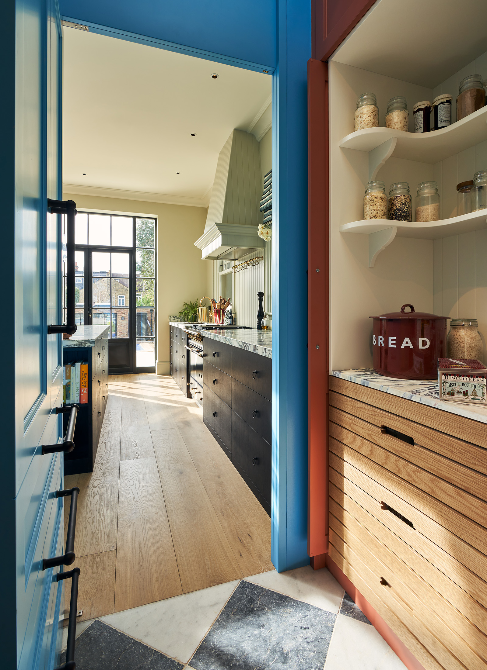 In the foreground is a kitchen pantry with open shelving displaying kitchen jars, marble worktops, wooden drawers and a checkerboard floor, and in the background is a classic kitchen with green-neutral walls, black cabinets and wooden flooring