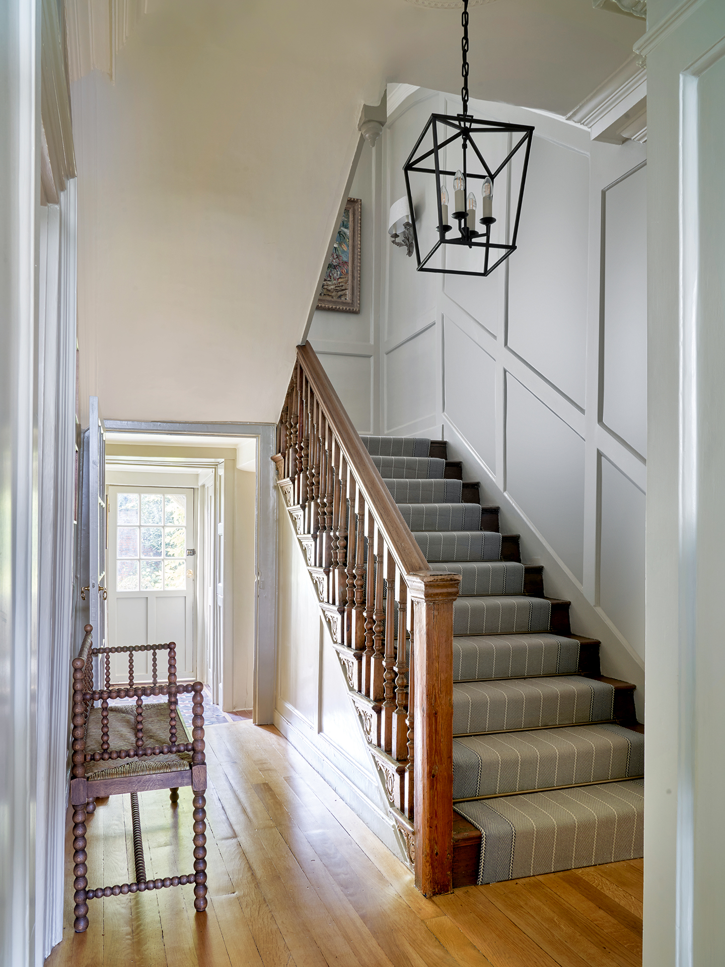 A grey panelled stairway with a striped runner on the stairs and wooden floorboards in the hall. A bobbin bench sits to the left and a large lantern-style pendant hangs above