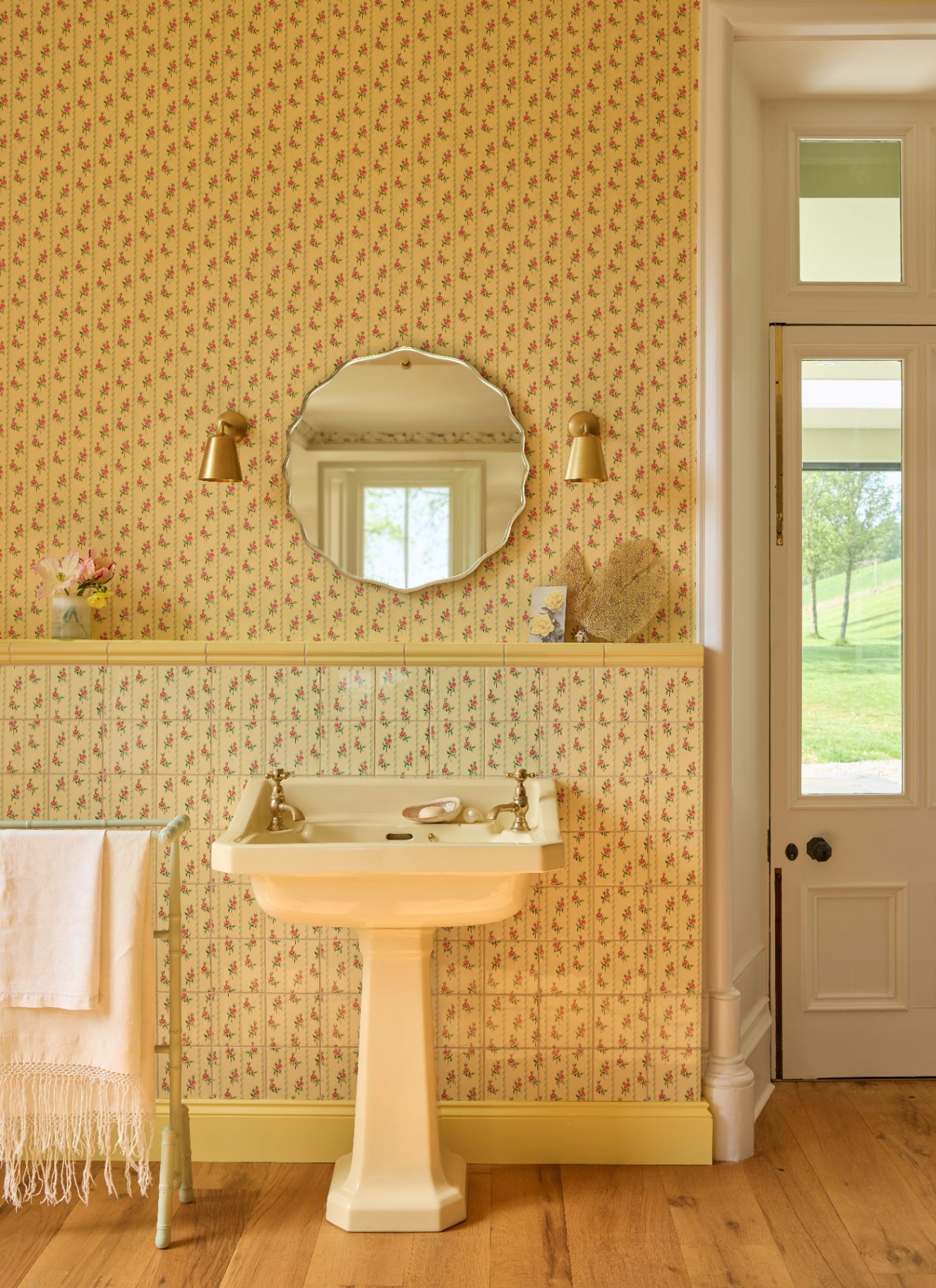 Pale yellow bathroom with a pedestal sink, scalloped round mirror, and brass sconces against floral wallpaper and matching tiled wainscoting. Wooden floors and a tall glass panel door complete the bright space