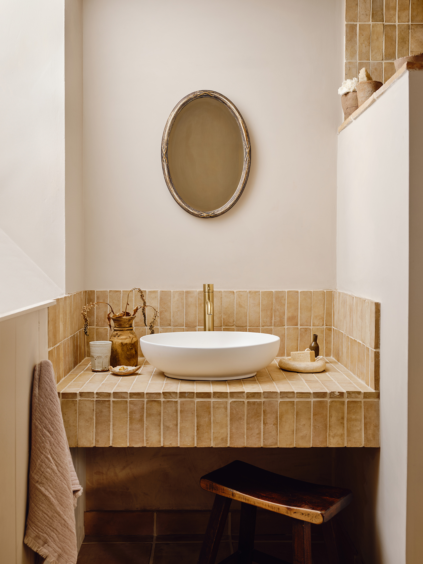 A close up of a built-in tiled shelf holding a basin in a bathroom. A wooden stool sits underneath, a towel hangs to the left and above is an oval mirror. 