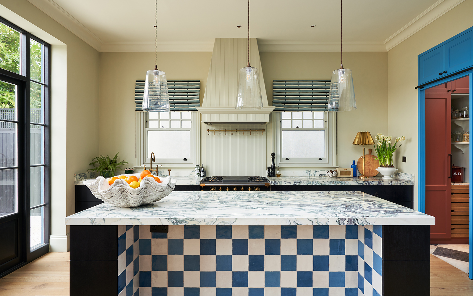 Kitchen with neutral walls and black cabinetry, in the centre is an island with blue and white checkerboard tiles in the alcove, the worktops are all in white veined marble and a large decorative clam shell filled with oranges sits on the island