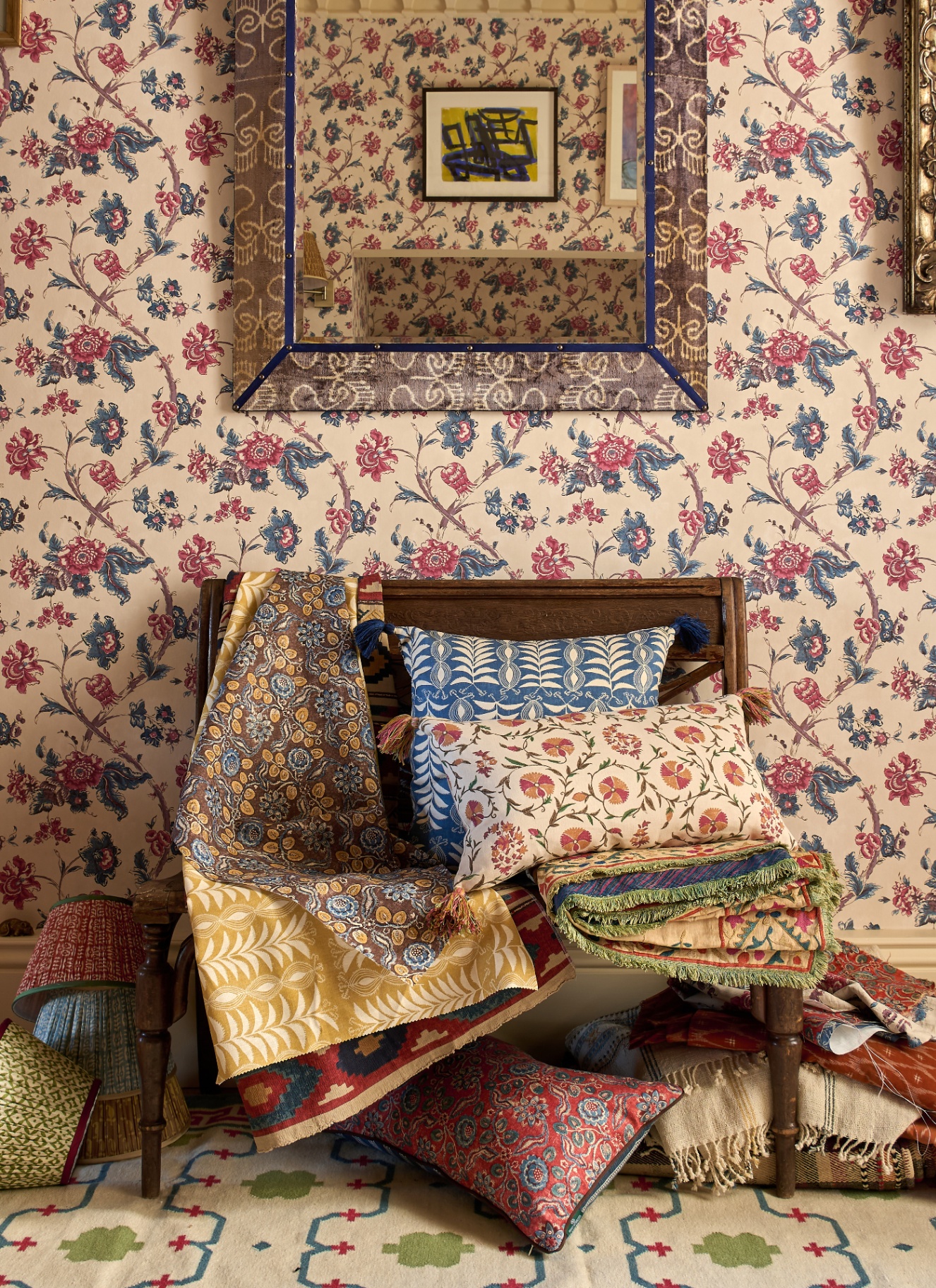 a pile of botanical patterned fabrics and cushions on a wooden chair against a cream, red and blue botanical wallpaper