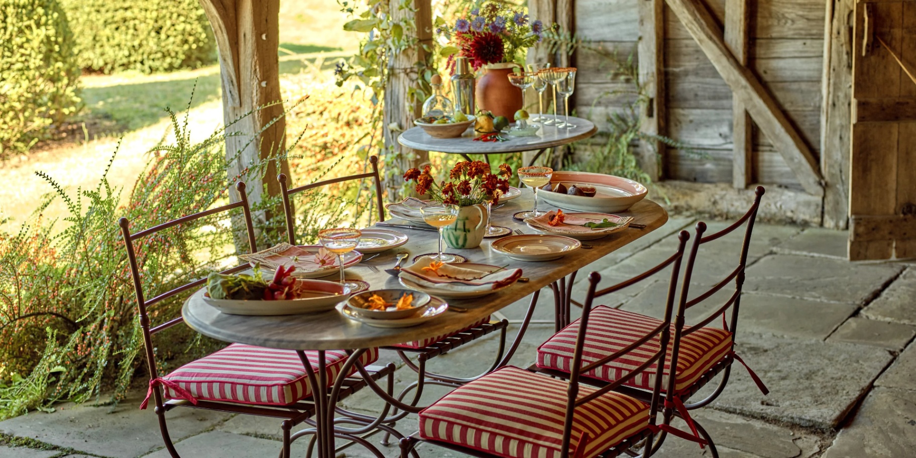 an outdoor dining table under a wooden roof, with 4 metal chairs with red and white striped seat pads and a table full of plates and food