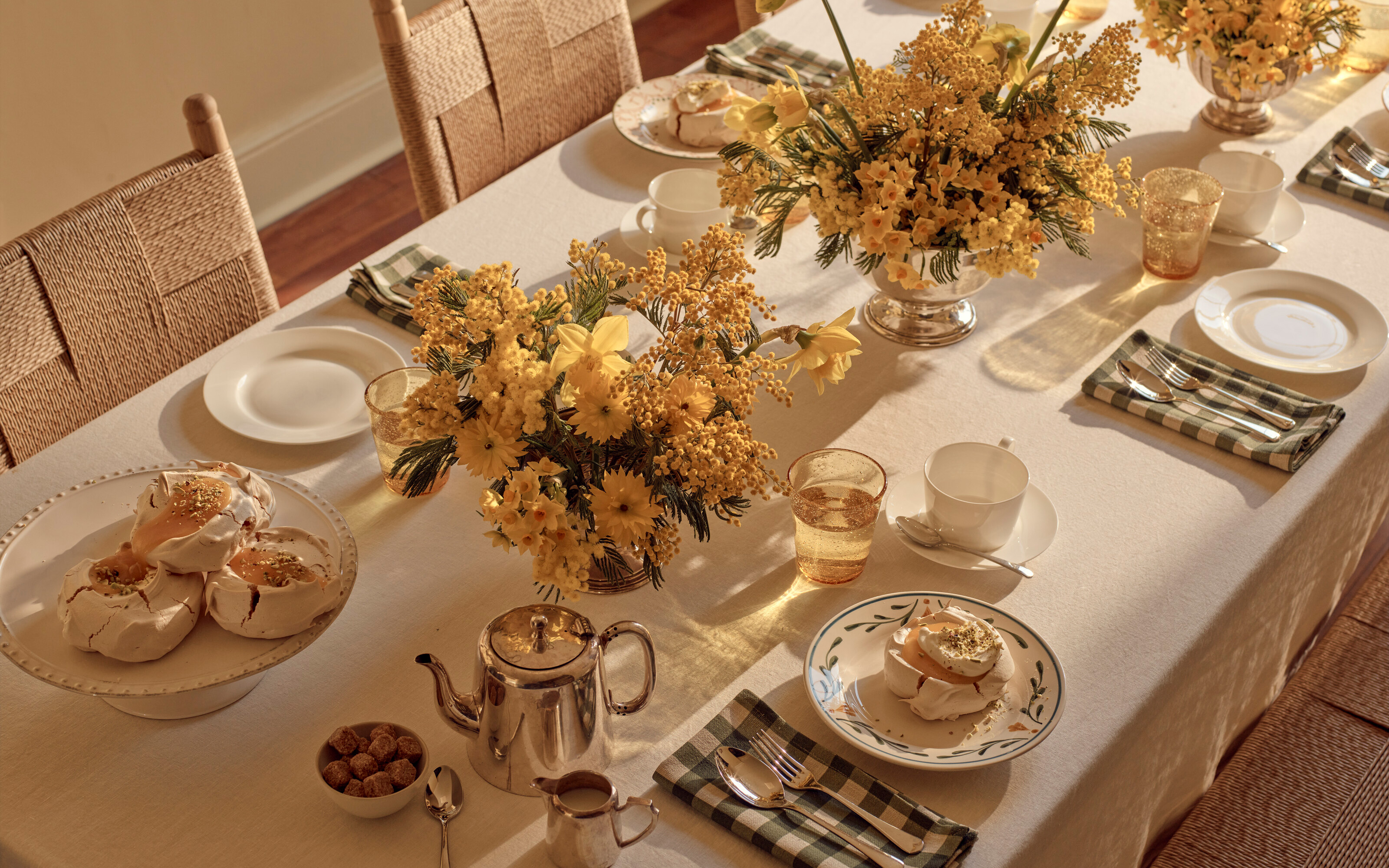 a tablescape with a cream table cloth, large silver vases in the middle of the table filled with yellow flowers and plates with green and cream gingham print
