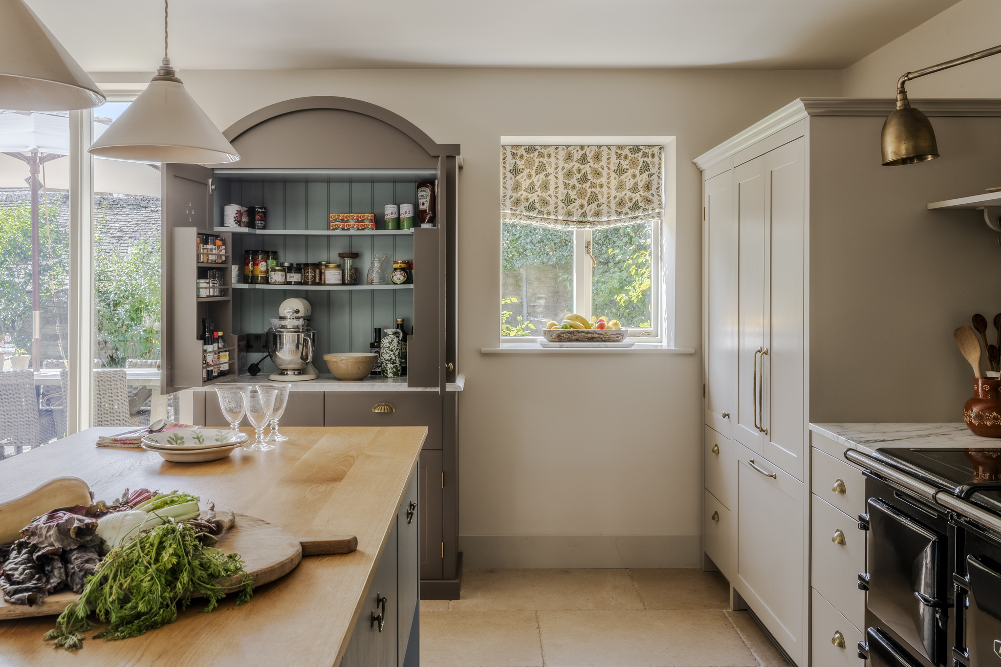 a country kitchen with an open pantry cupboard full of spices and a stand mixer and a farmhouse table with vegetables on