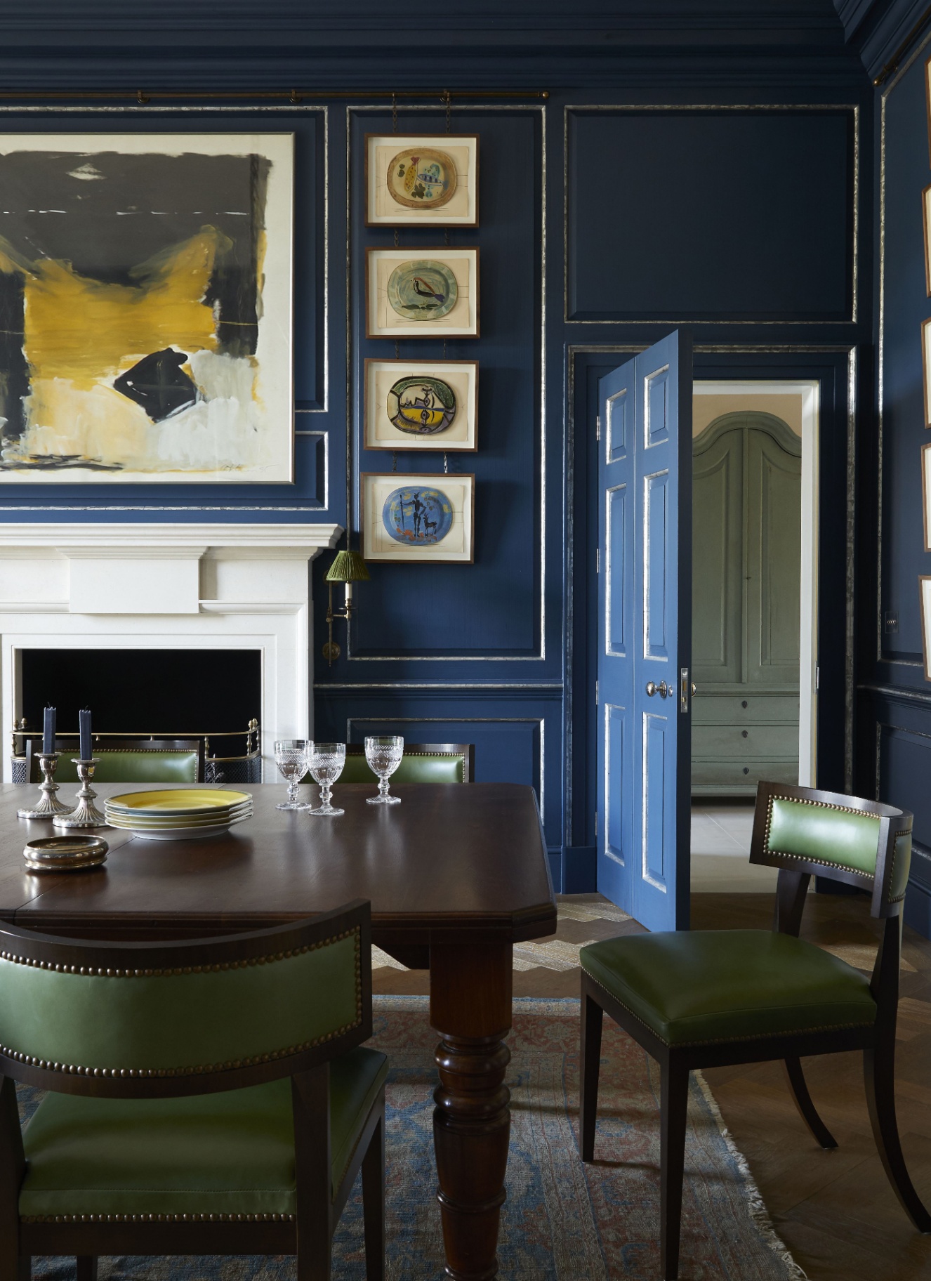 Dark navy blue painted dining room with gold framed artwork, a dark wood dining table set with crystal glassware, green leather dining chairs, and a glimpses of an adjoining room through an open blue door
