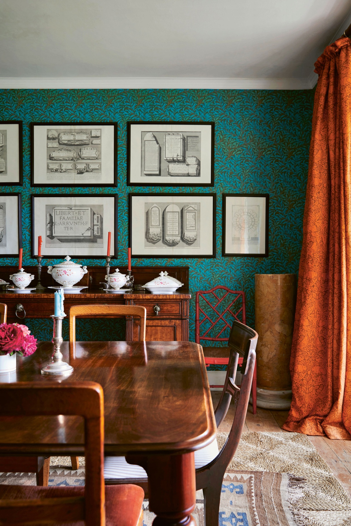 a dining room decorated with a blue and green botanical wallpaper, with framed drawings displayed on top. In the foreground is a wooden dining table and chairs atop two layered rugs and to the right, an orange curtain hangs