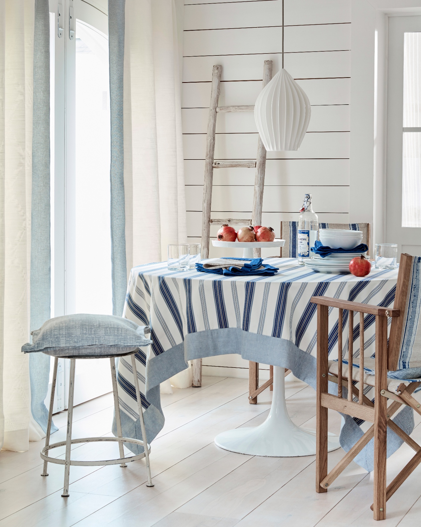 A dining room with a round table covered in a blue and white stripe table cloth, two dining chairs and a stool with blue cushions, a wooden ladder leaning on the wall and a large white pendant above the table